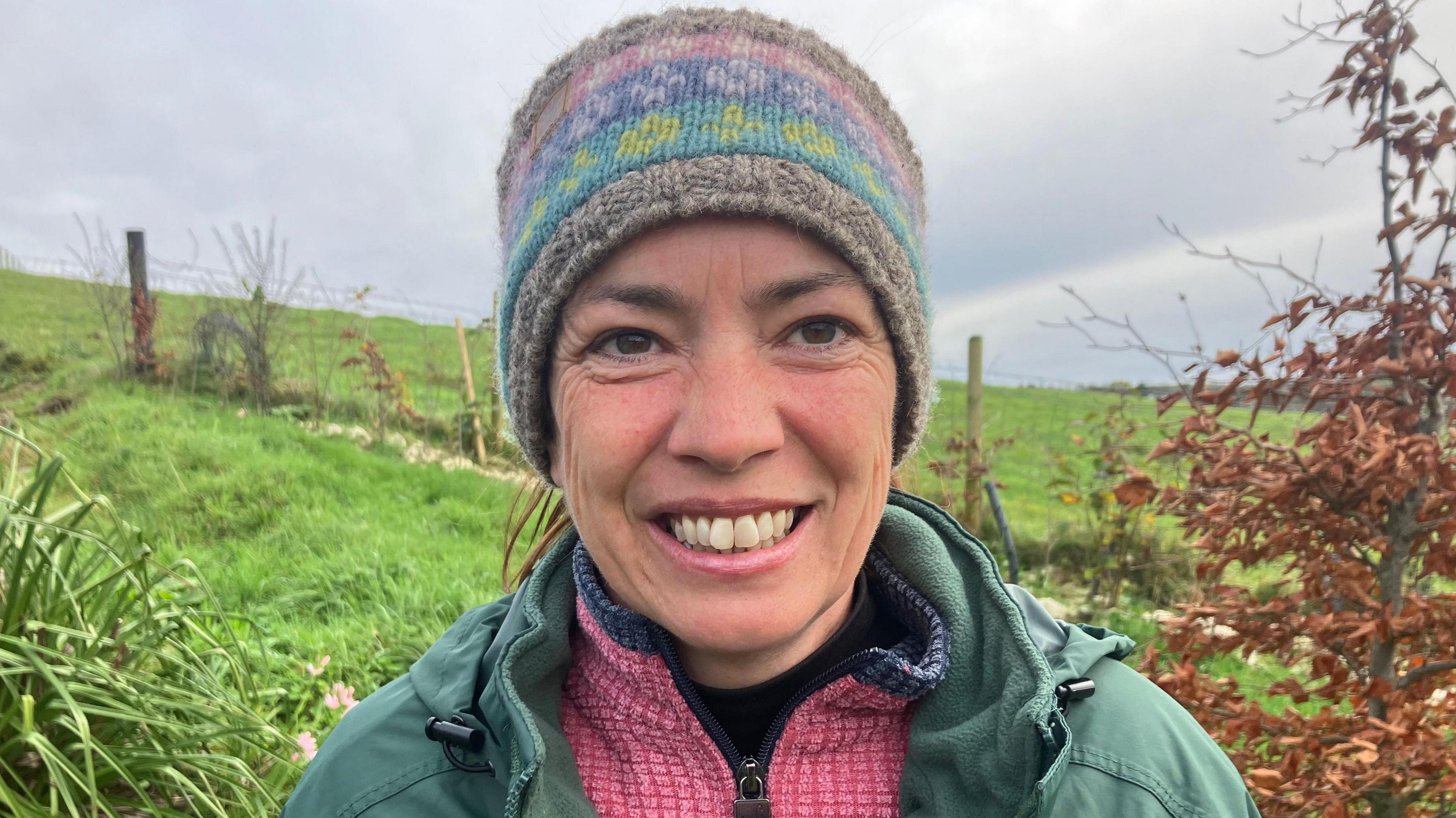 A photo of Anne Brown who is wearing a head warmer with a grey, blue, yellow, dark blue, pink and grey lined pattern. She is wearing a pink fleece with a green coat over it. She is standing outside by a grass field. 