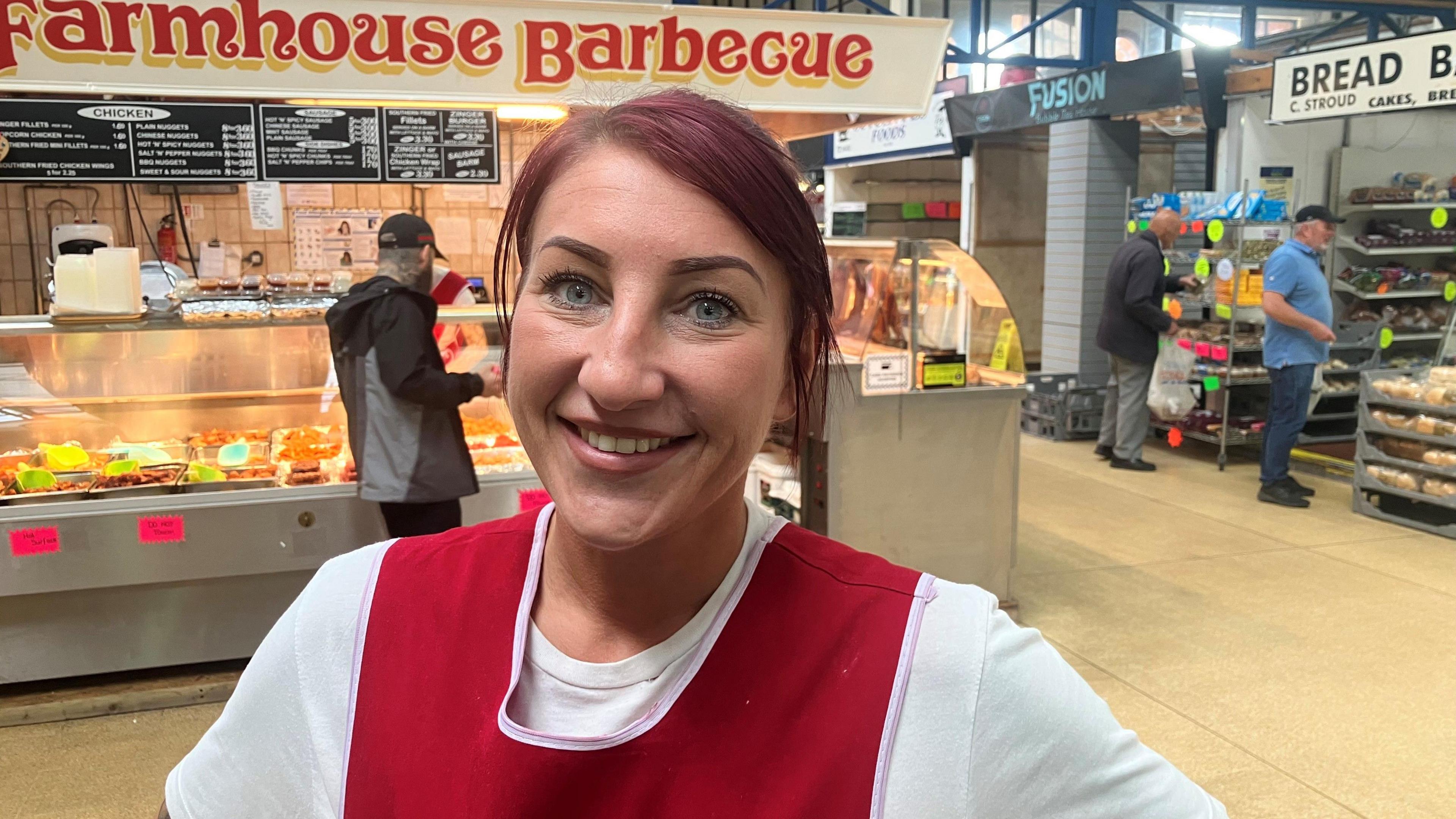 Photograph of Laura Tomlinson who works at Wigan indoor market. Wearing a white top with a red apron over the top, she is pictured in front of June's Farmhouse Barbecue stall.