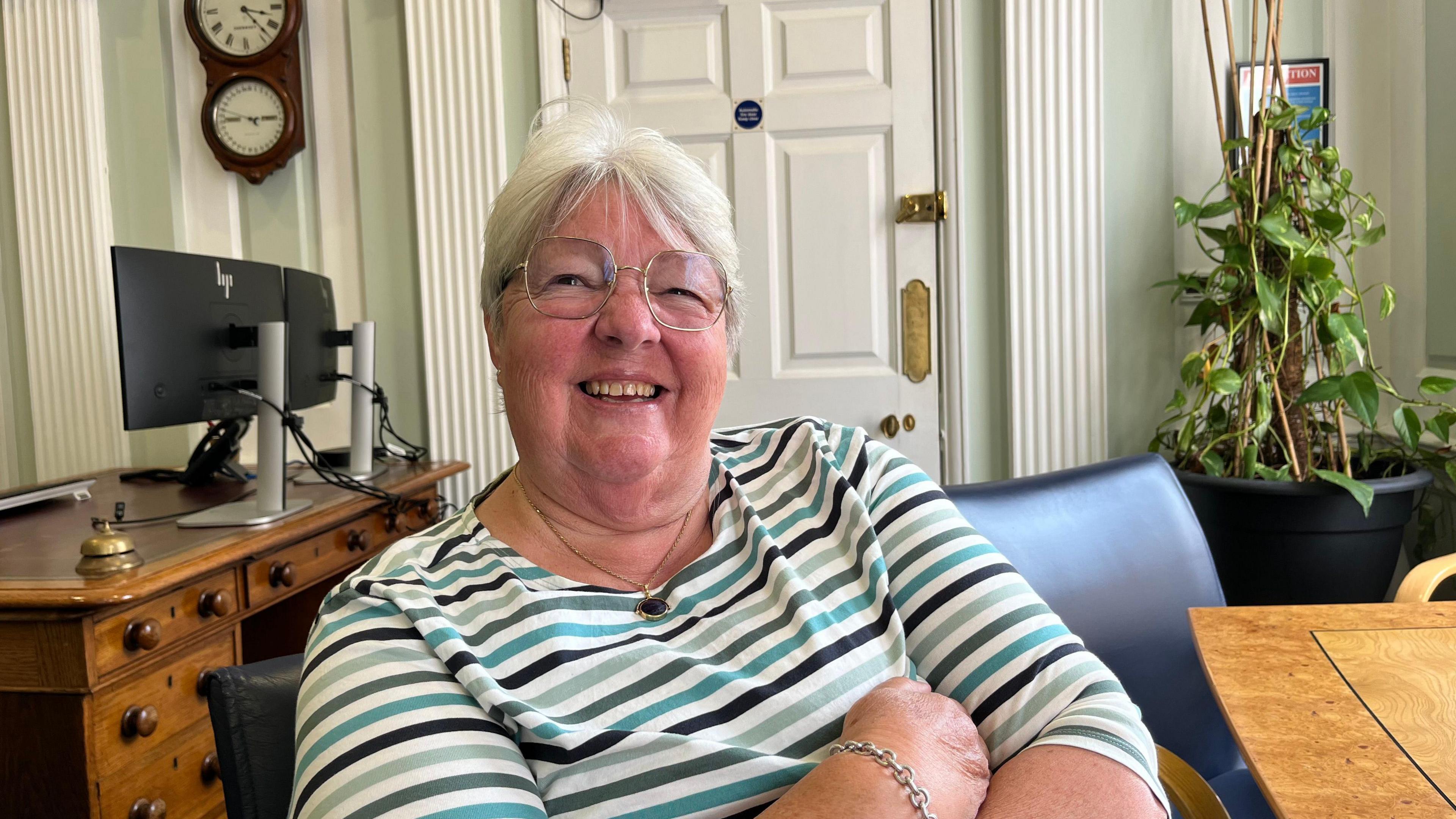 A woman in a striped T-shirt and glasses sitting in an office and smiling at the camera