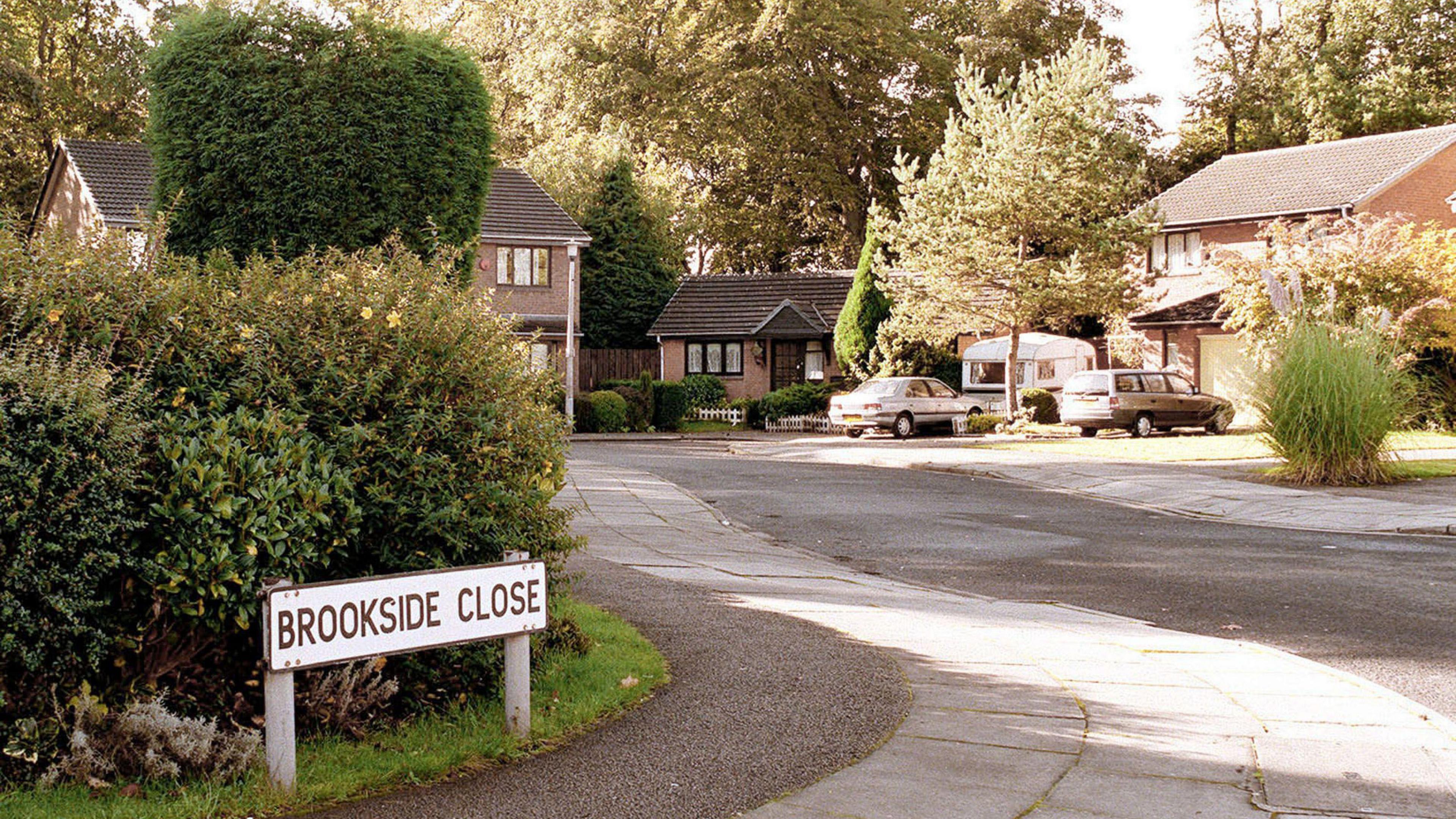 A picture of Brookside Close, captured in the 1990s. To the left is a street sign which reads: Brookside Close with bushes behind it. The road curves round to the left with a number of houses and one bungalow in view.