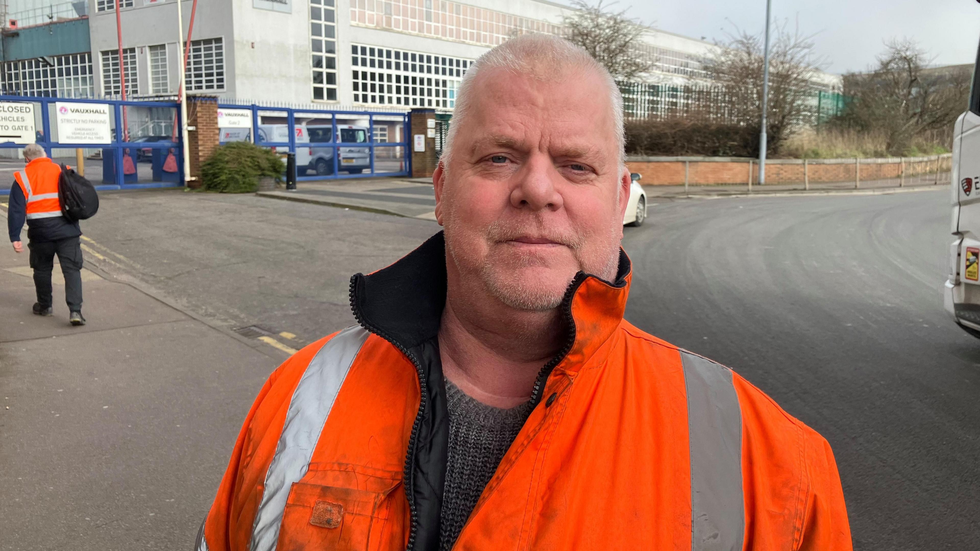 Peter wears a bright orange high vis coat with reflective grey strips, open at the neck. He has a grey jumper beneath it. He has white short cropped hair and stubble, and a half smile. The factory is behind him and another worker is seen from behind walking towards it.