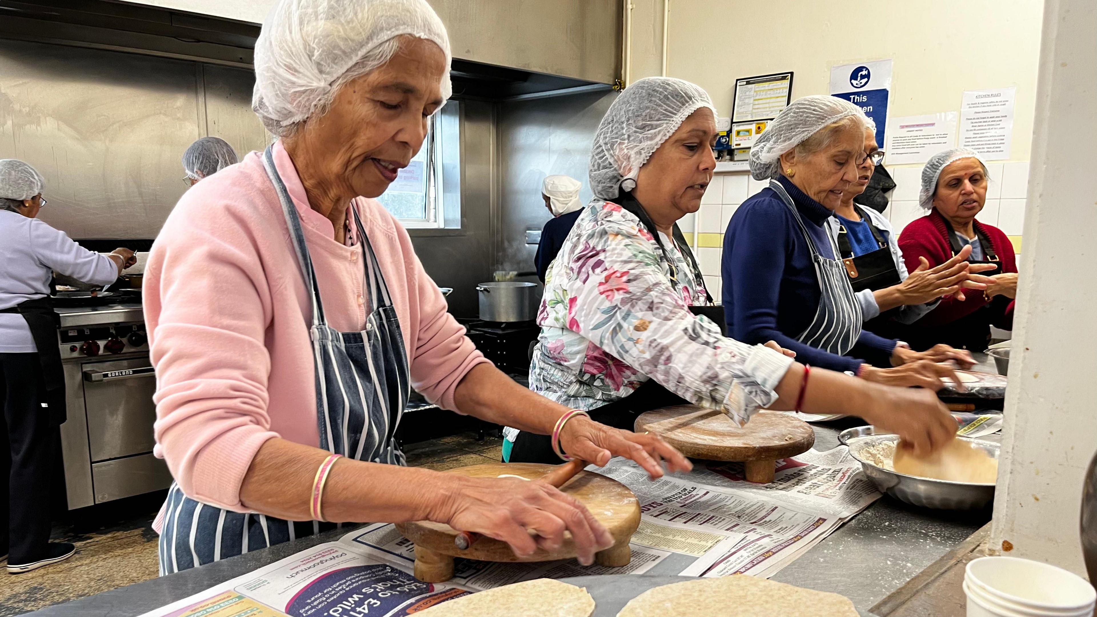 A row of women in the kitchen rolling dough to create rotis for the lunch club. All are wearing hair nets and aprons.
