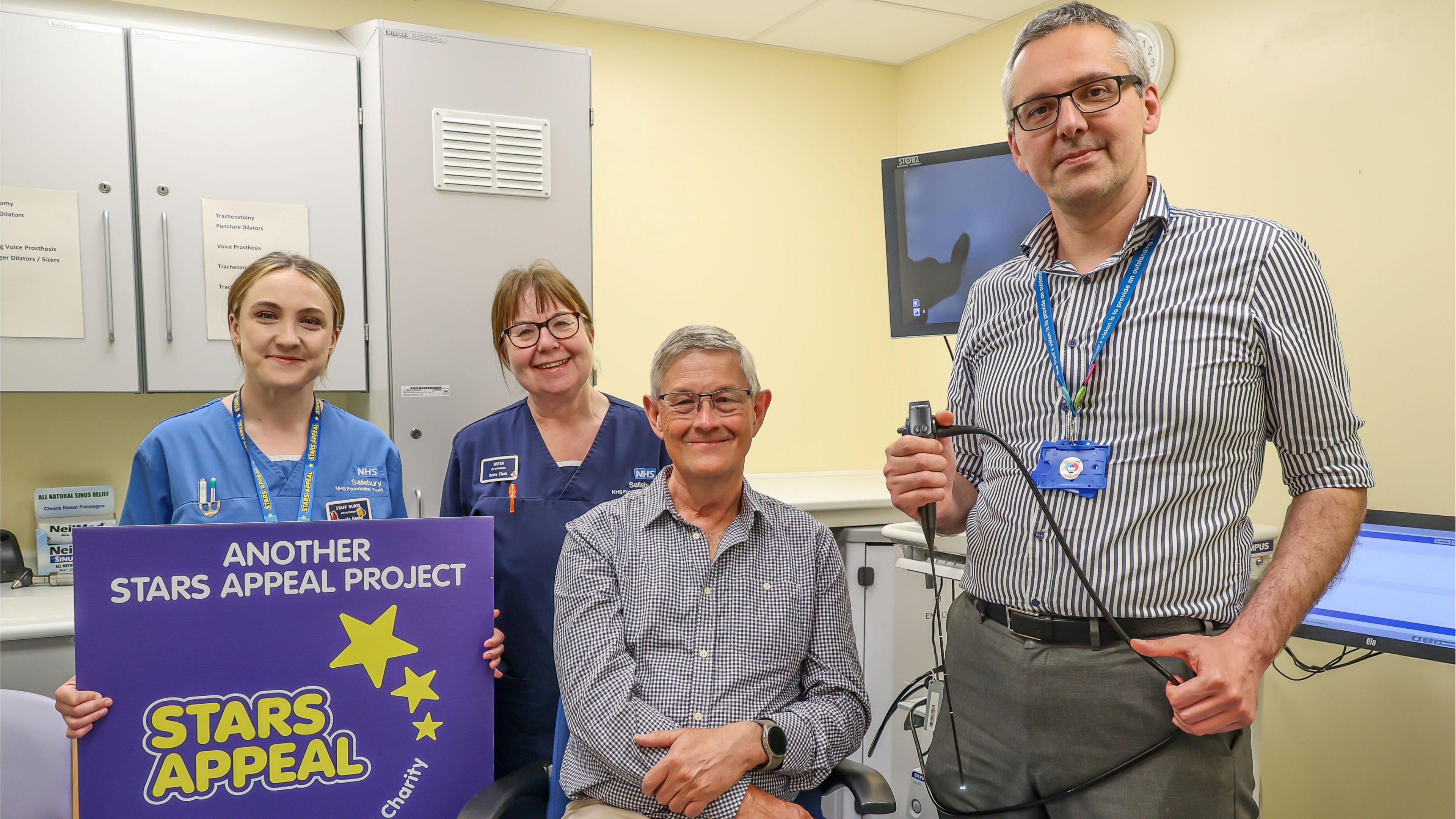 Peter Russel and Kostas Marinakis are in a hospital room with two female nurses behind them. One of the nurses is holding a purple sign which says 'another stars Appeal Project' and has the Stars Appeal logo.