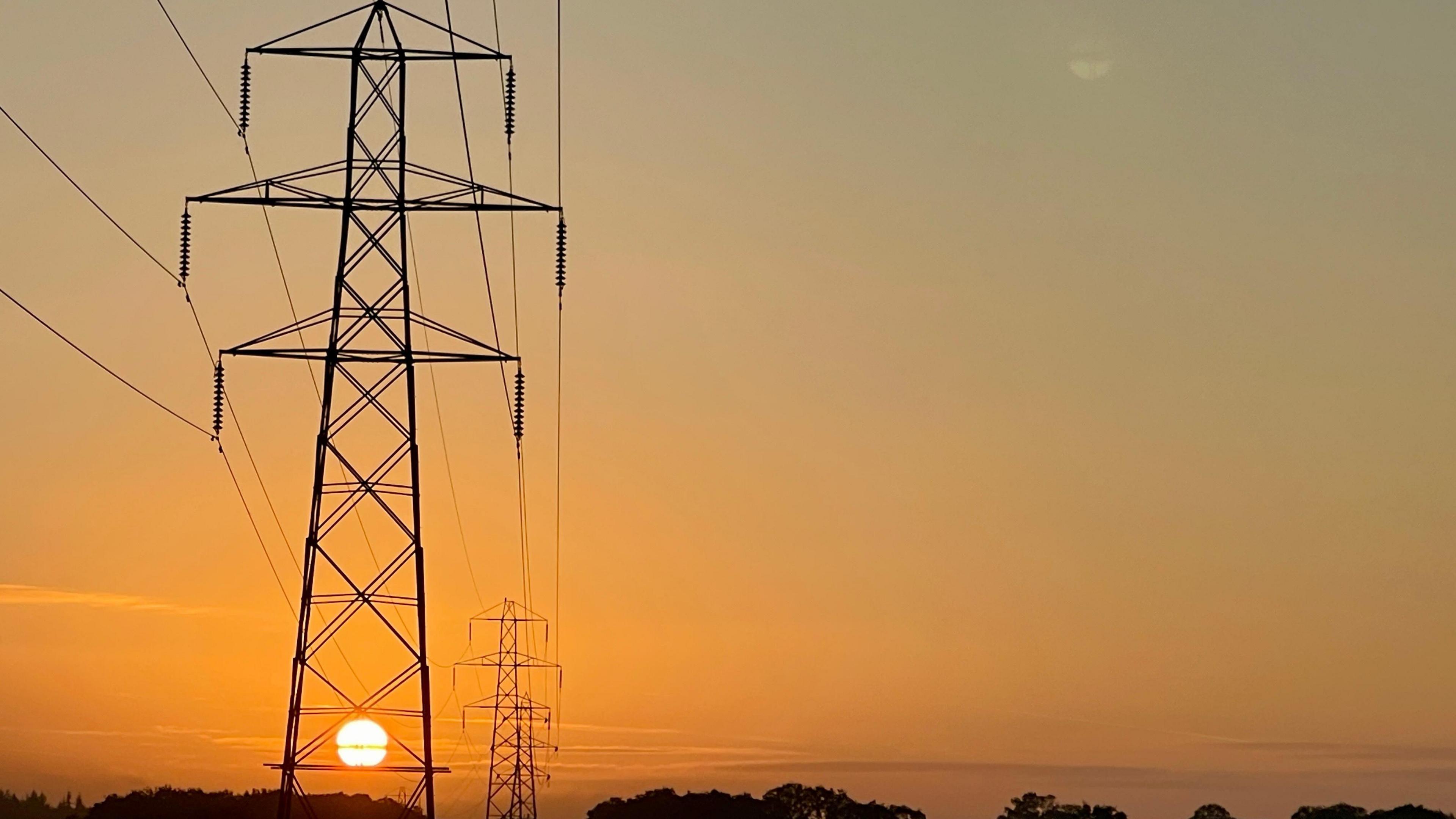 The rising sun shines orange through the metalwork of an electricity pylon which rise above the silhouette of trees on a low horizon. The wide expanse of sky is illuminated with a graduating orange emanating from the early morning sun.