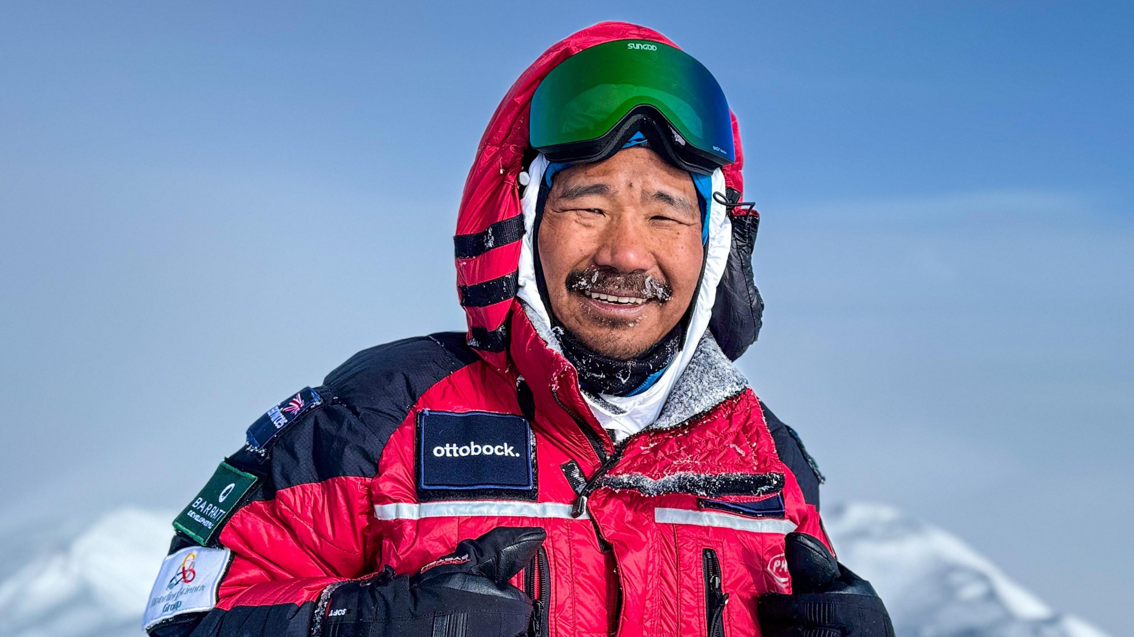 A man in a red snowsuit and a snow-coated beard smiles at the camera. There are snowy mountains in the backdrop. 