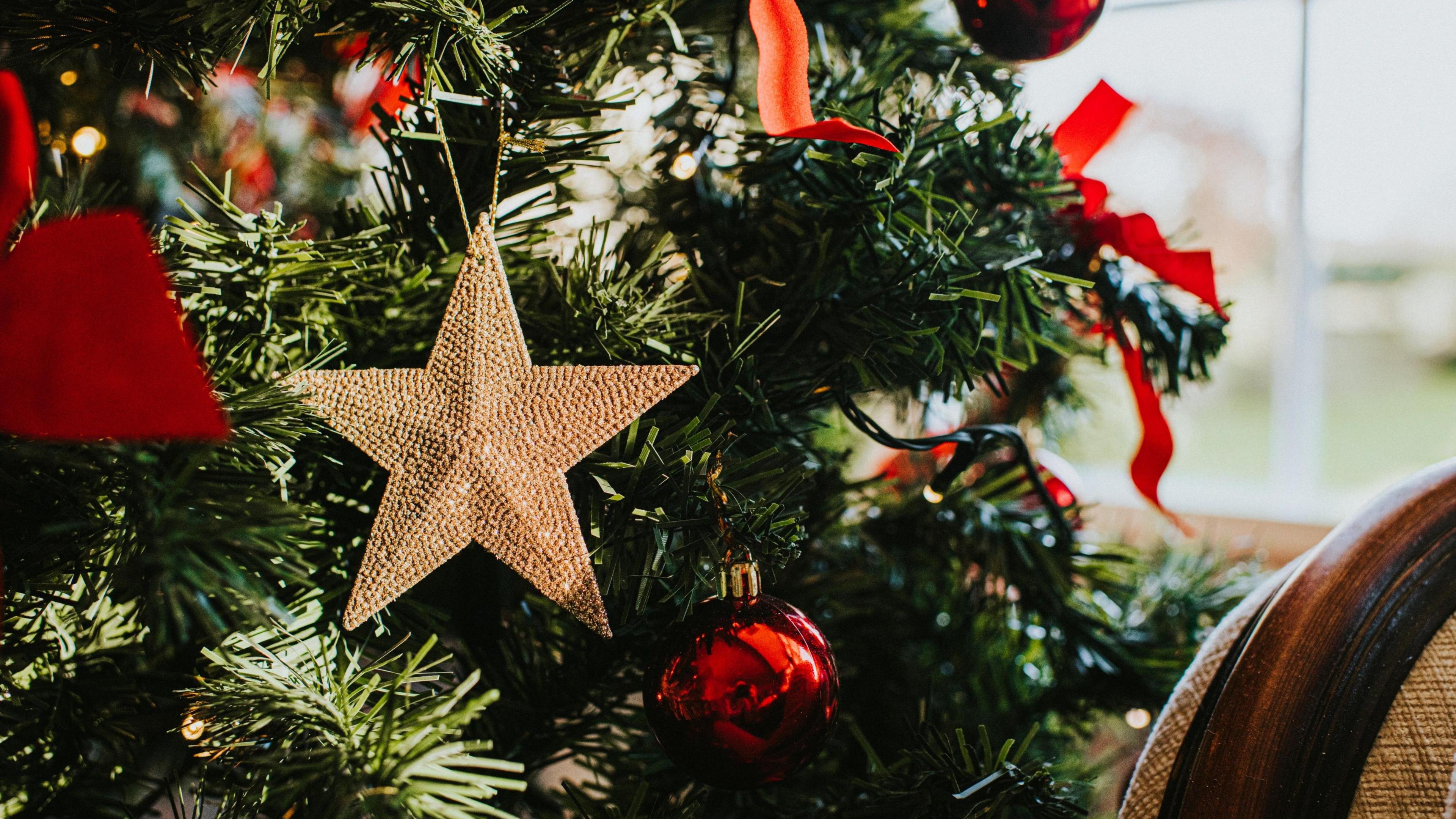 Ornamental gold star decoration hanging on a branch on an artificial Christmas Tree. Image focuses on the star but it is surrounded by red ribbons and baubles and other decorations.