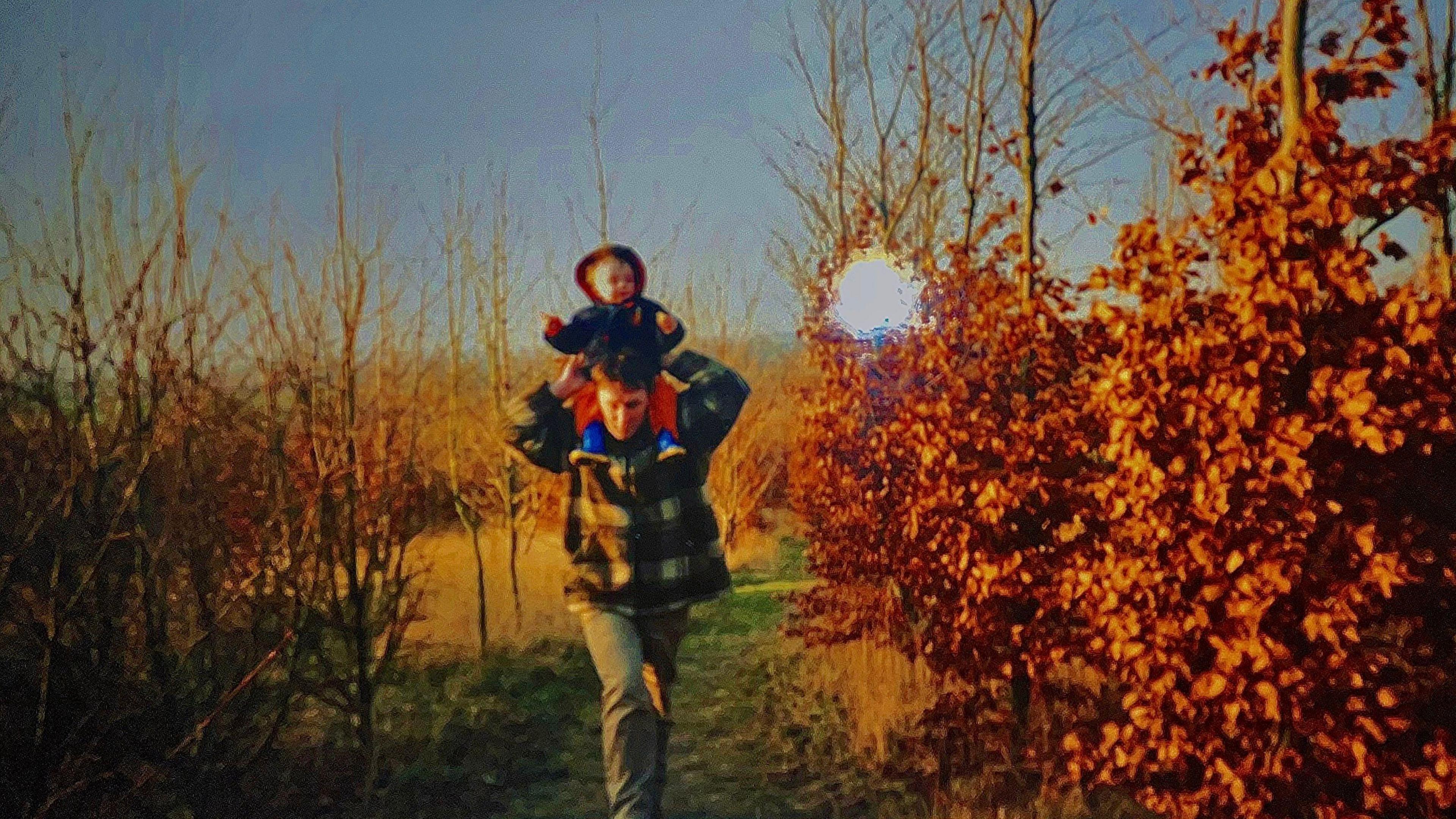 Man with green trousers holding a child on his shoulders on a path between brown-leaved trees. The sky is visible in the background. The picture was taken on an old camera, possibly a Polaroid