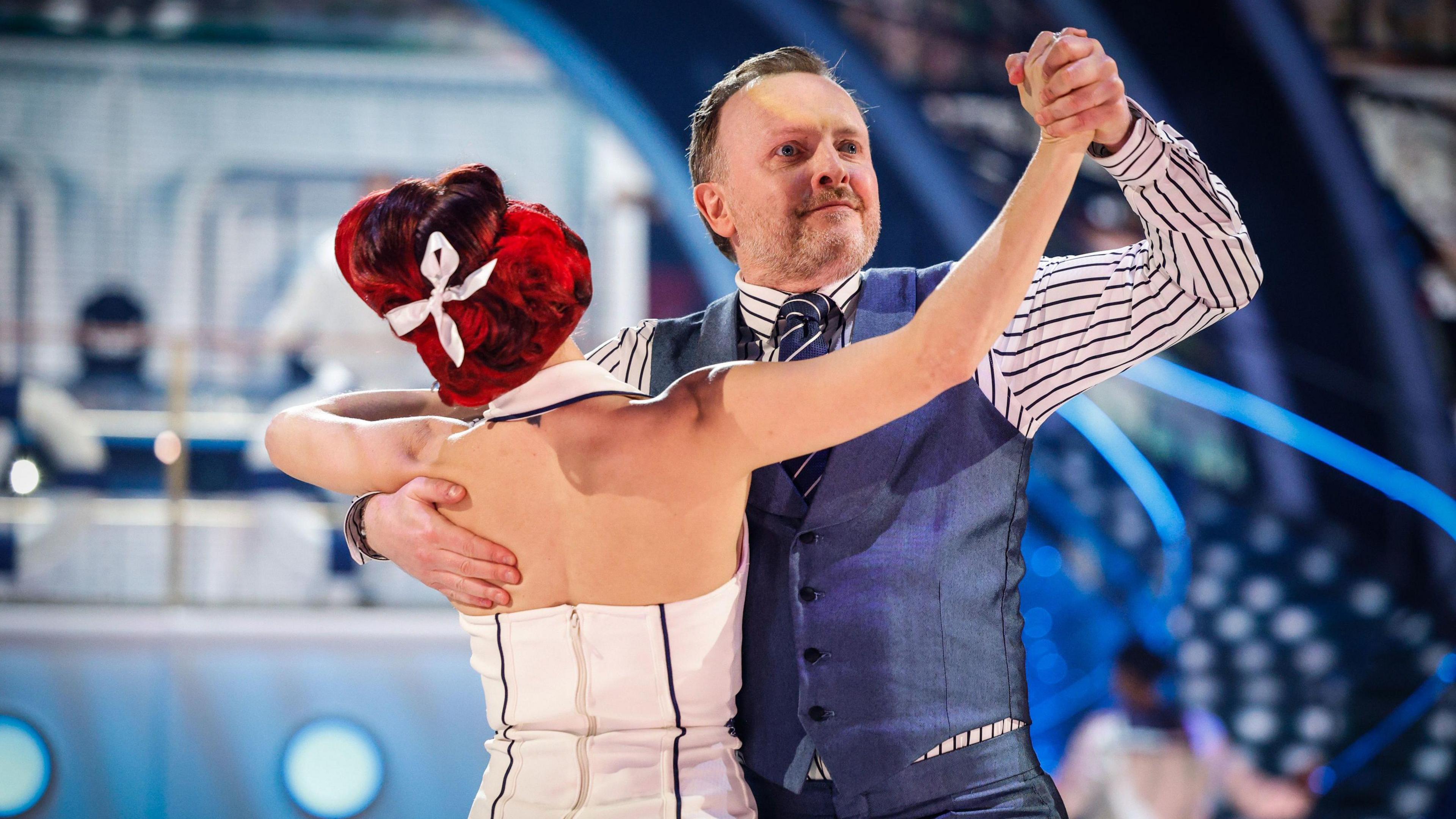 Comedian Chris McCausland dances with his partner Dianne Buswell. They are holding hands and facing each other in a waltz position. He is wearing a white shirt with purple stripes and a contrasting tie with purple waistcoat and trousers. Dianne Buswell has her back to the camera and wears a white dress and has a white bow tied in her red hair.