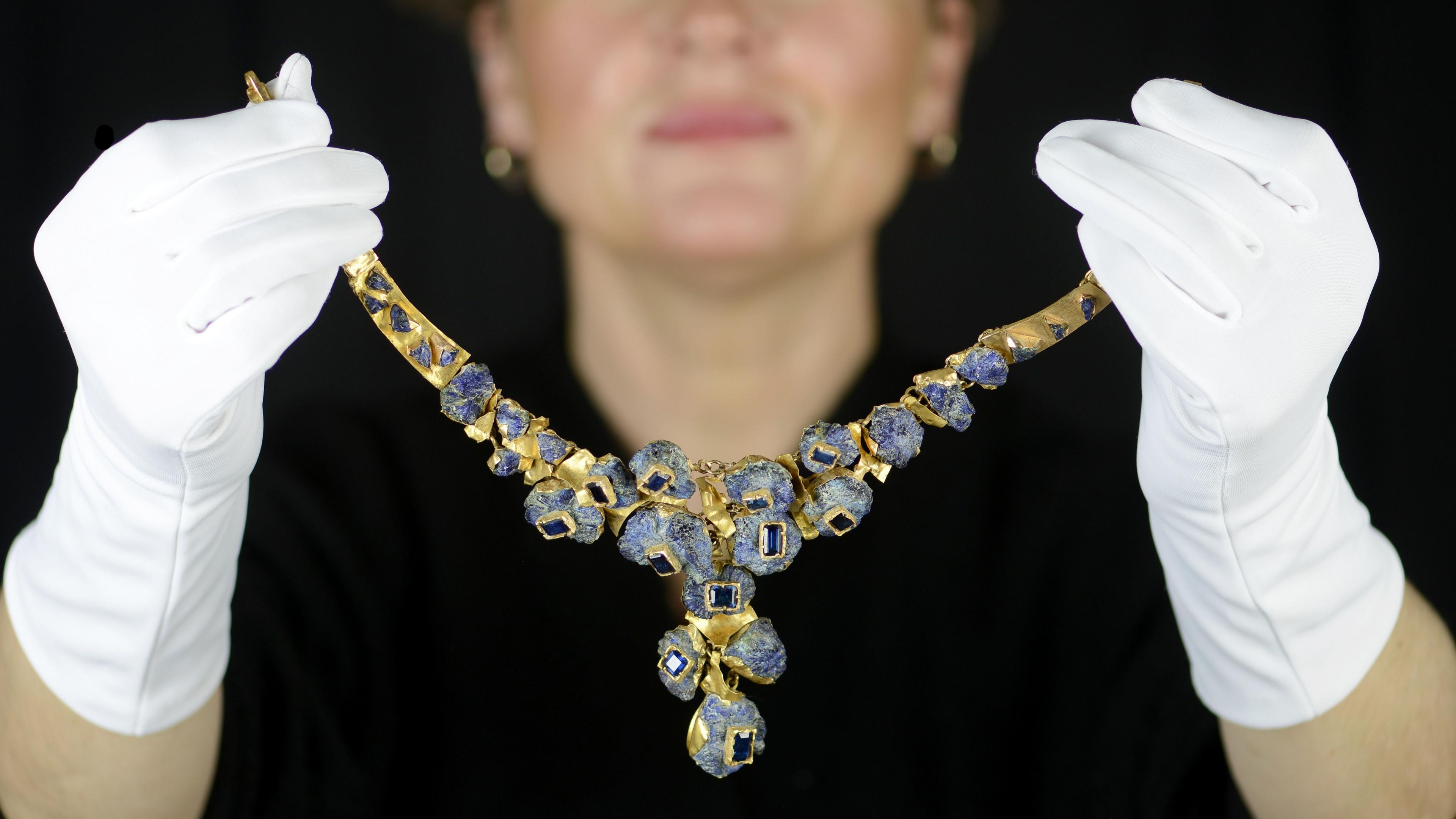 A women with white gloves on holding a large gold and blue rock like necklace, with square blue gems in each connecting link.