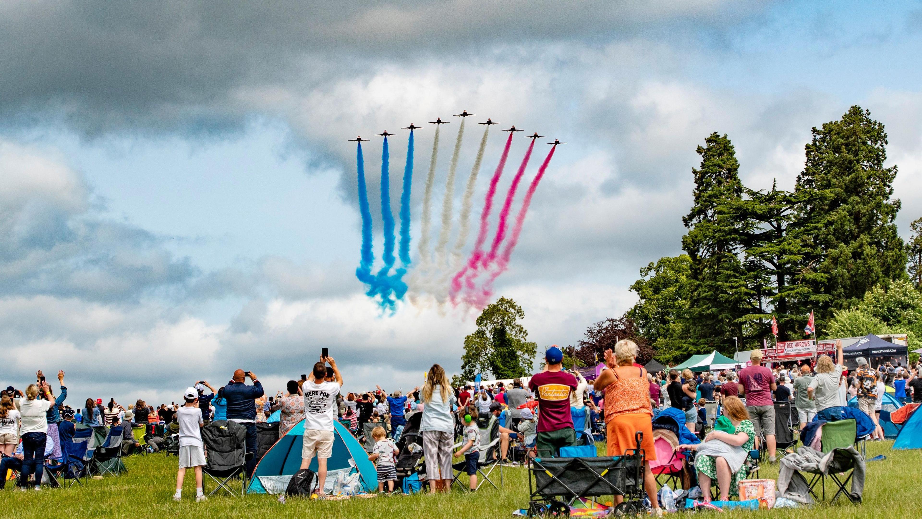 Nine RAF Red Arrows fly over crowds who are taking pictures of the aircraft with blue, white and red smoke streaking across the sky