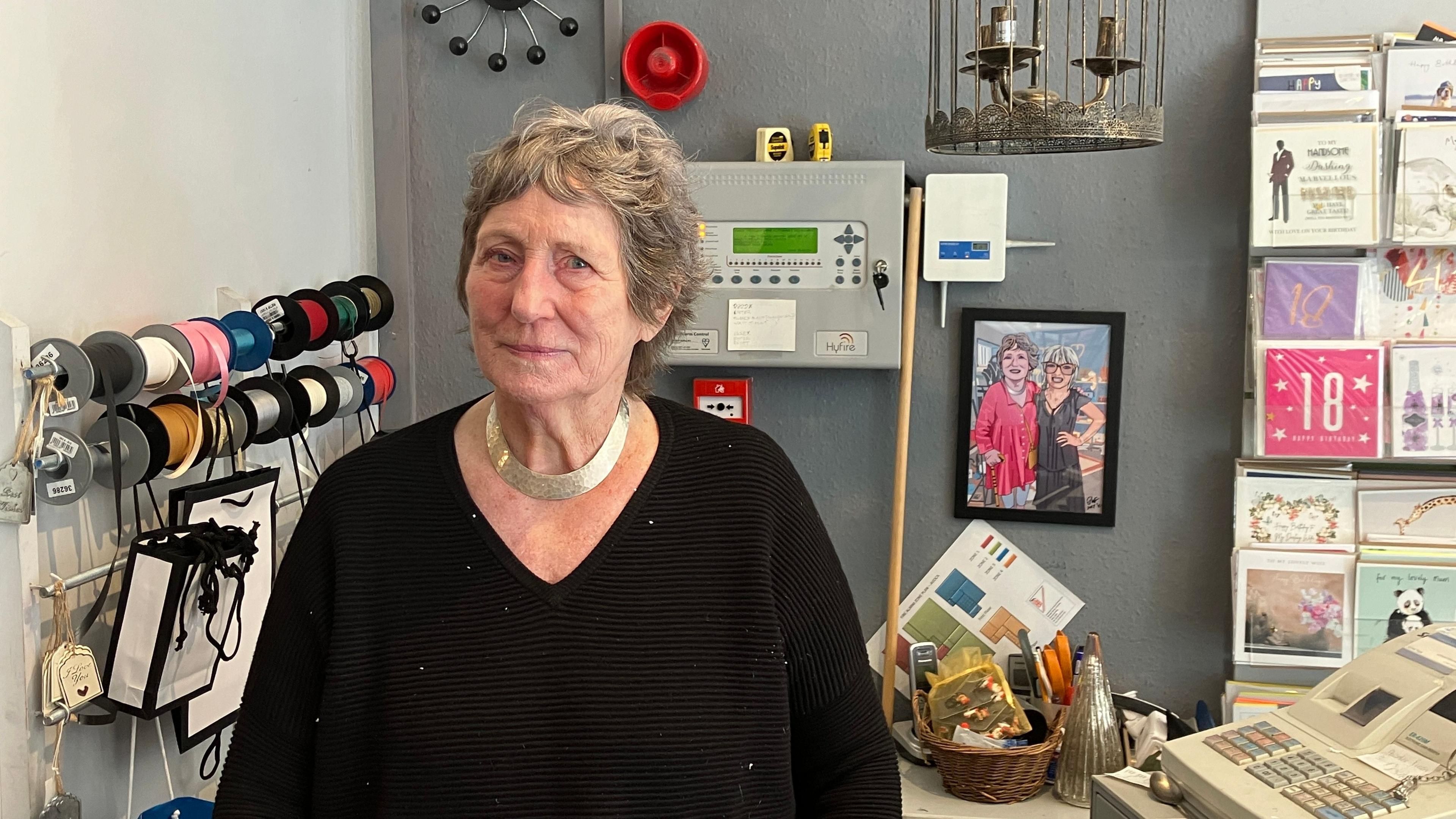 Maureen with short grey hair wearing a black jumper standing behind the till of her gift shop 