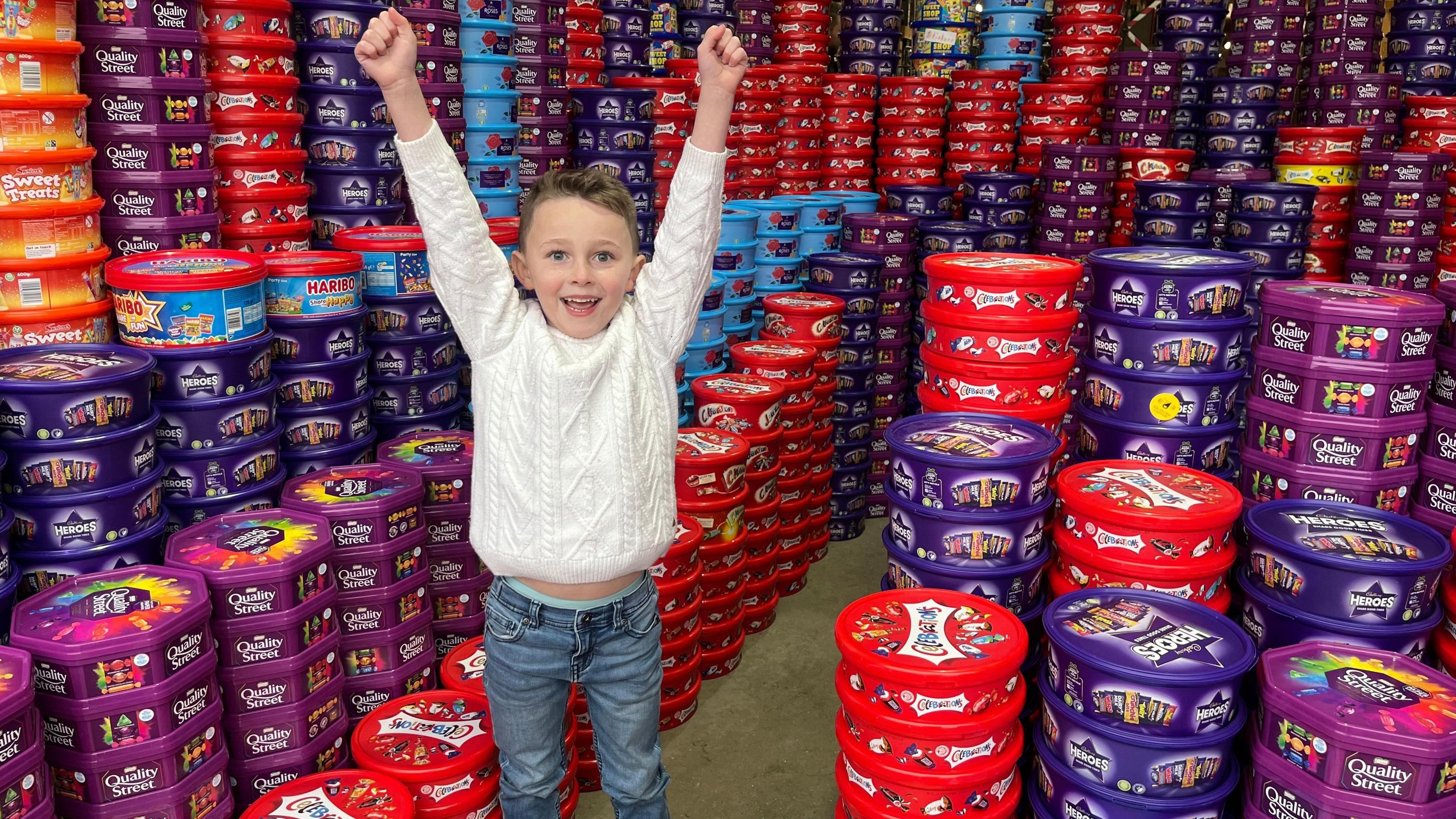 Teddy wears a white jumper and jeans. He has short light brown hair. His arms are raised in triumph. He is in a warehouse full of thousands of sweet tubs, including Heroes, Quality Street, and Celebrations.