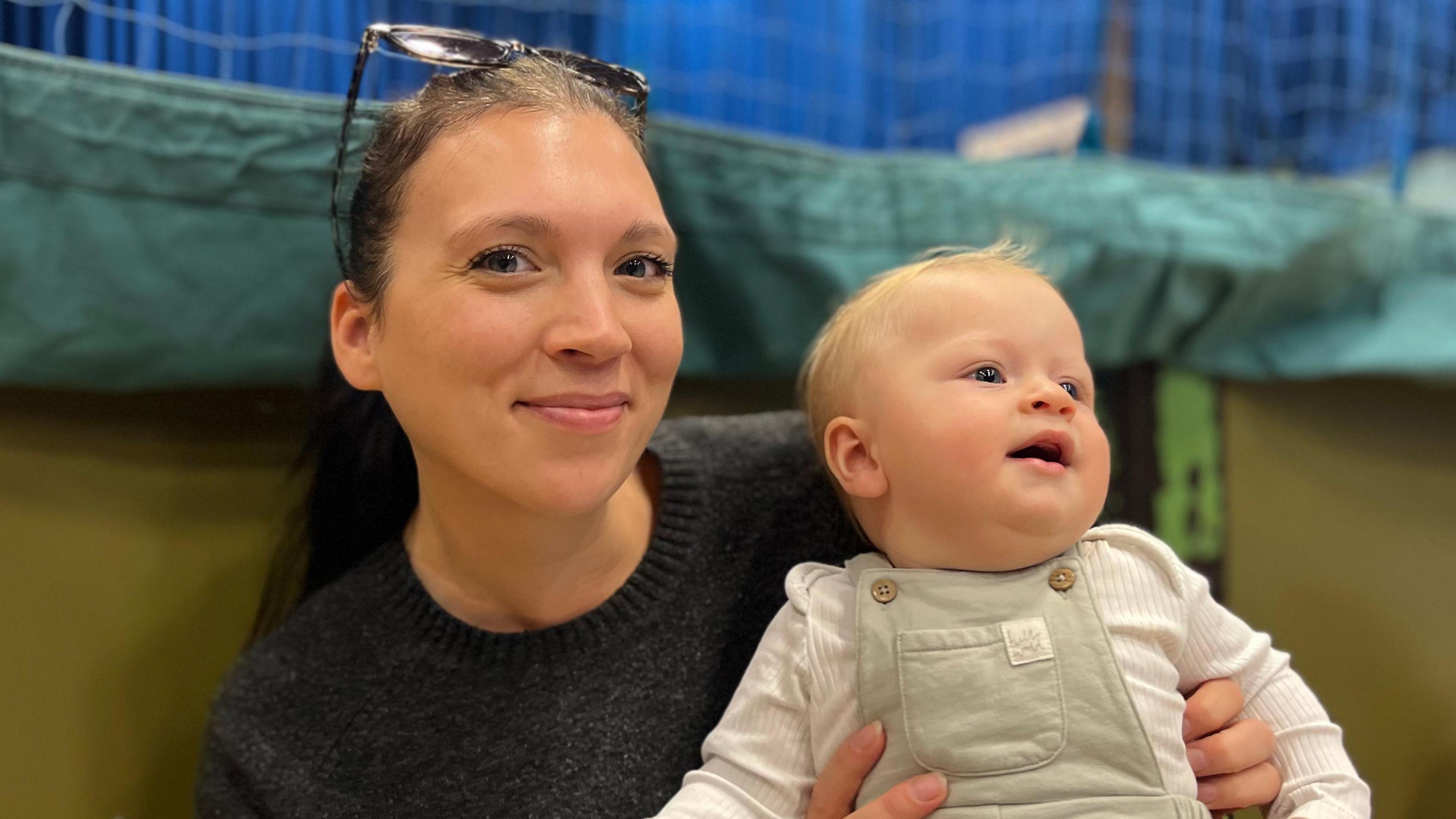 Leigh Walker and her baby Clyde - A woman with sunglasses on her head smiling at the camera. She's black hair in a ponytail and a black jumper. She is holding on her lap her baby who is wearing green dungarees and a white baby-gro. He's looking away from the camera and has a tuft of blonde hair on his wee head. 