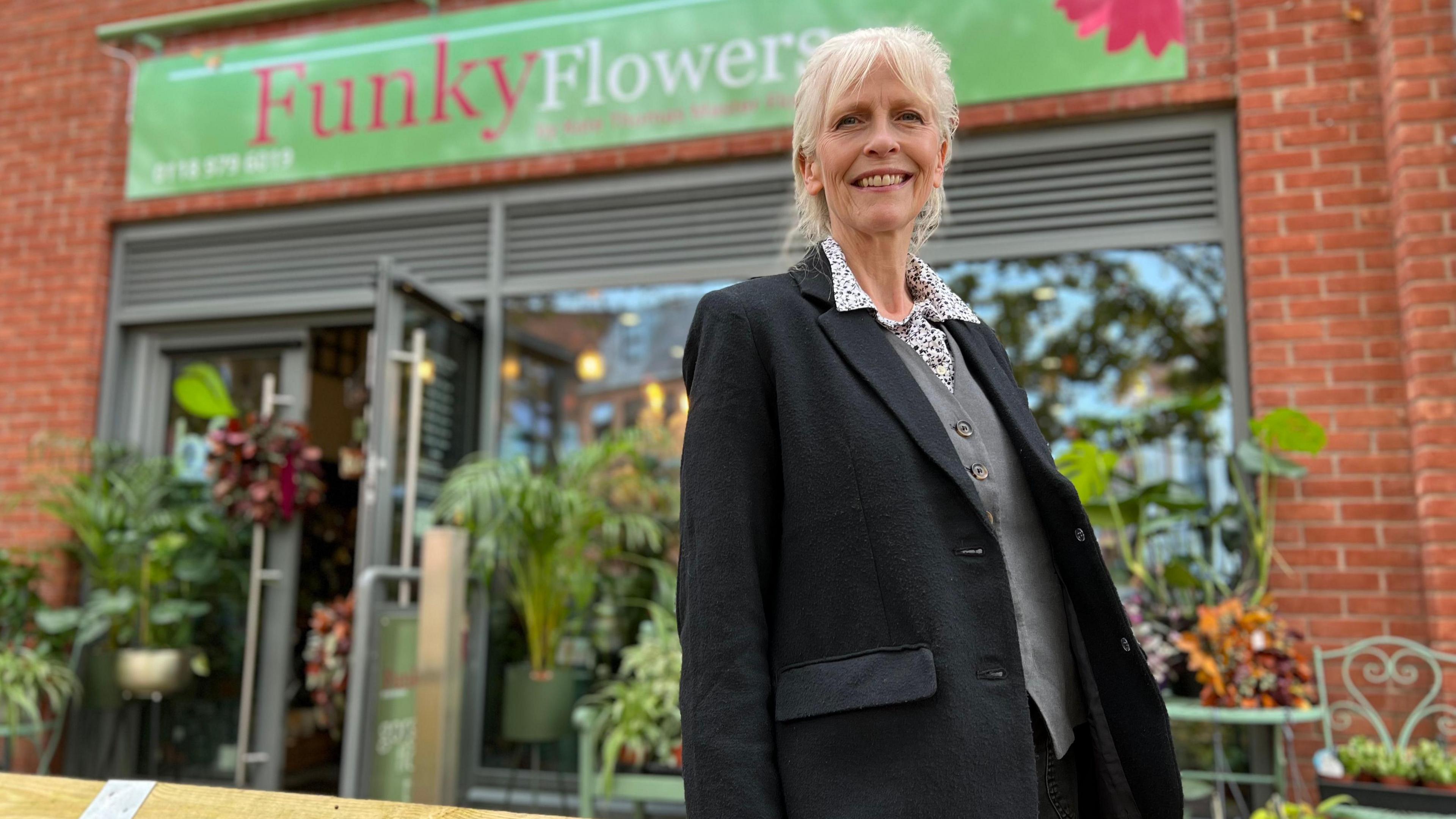 Kate Thomas stands outside her store, Funky Flowers, she is looking at the camera and smiling, her long white hair is tied back in a plait and she has a fringe framing her face. Kate wears a black blazer, with a whit eand black polka dot blouse and grey waistcoat with wooden buttons done up, underneath. Her store is slightly blurred behind her, the Funky Flowers sign is above the doorway, it is a green banner, with the word Funky in pink and Flowers in white, there are plants lining the entryway to the shop.