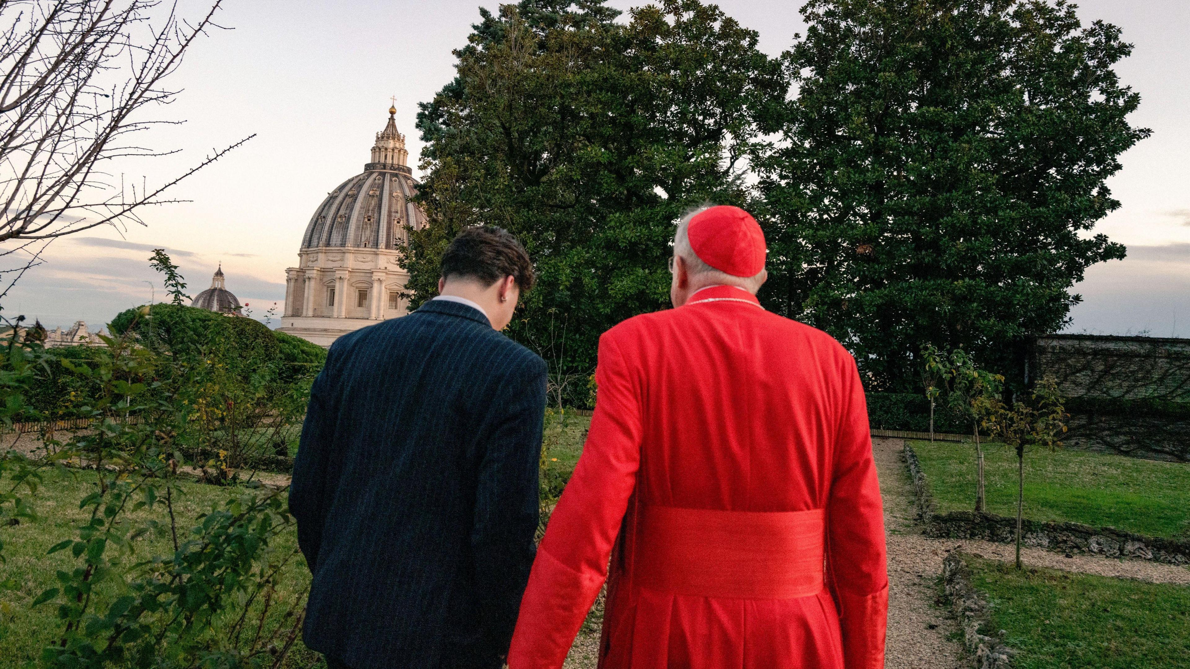 A man in a black suit walks with a Cardinal wearing red in the gardens of the Vatican.
