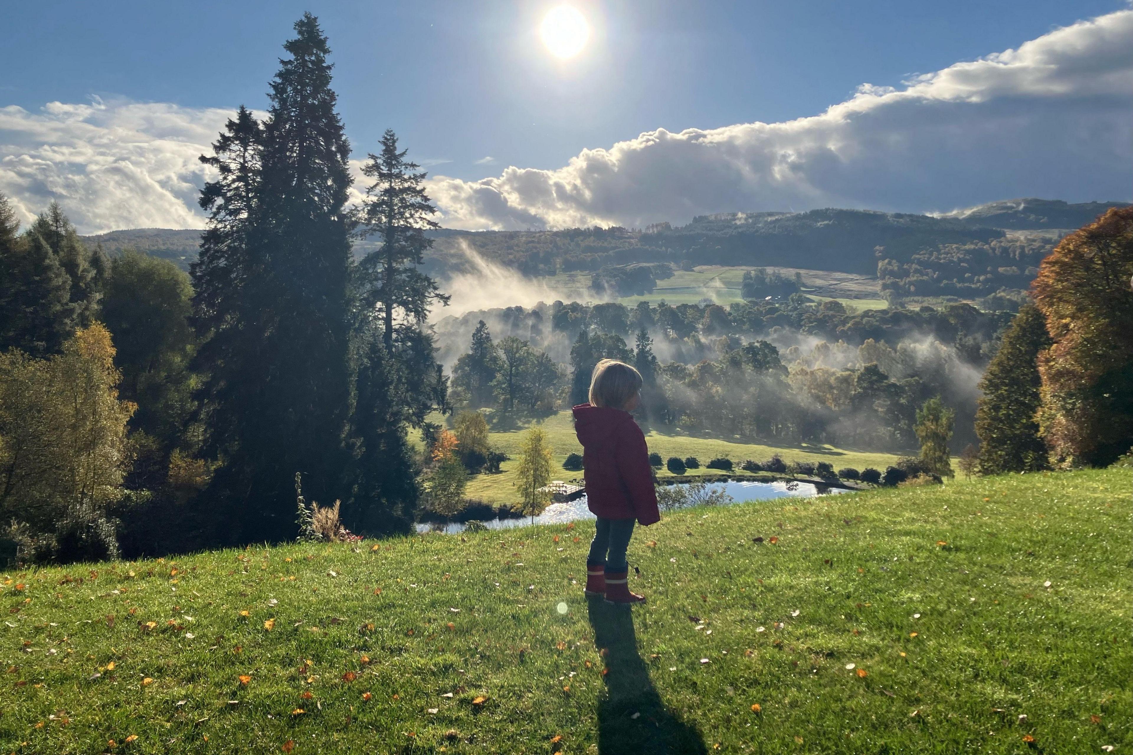 A boy in a red coat and welly boots stands looking into a misty valley with his back to the camera. The sun is high in the sky and there are dark green trees on the left hand side of the valley.