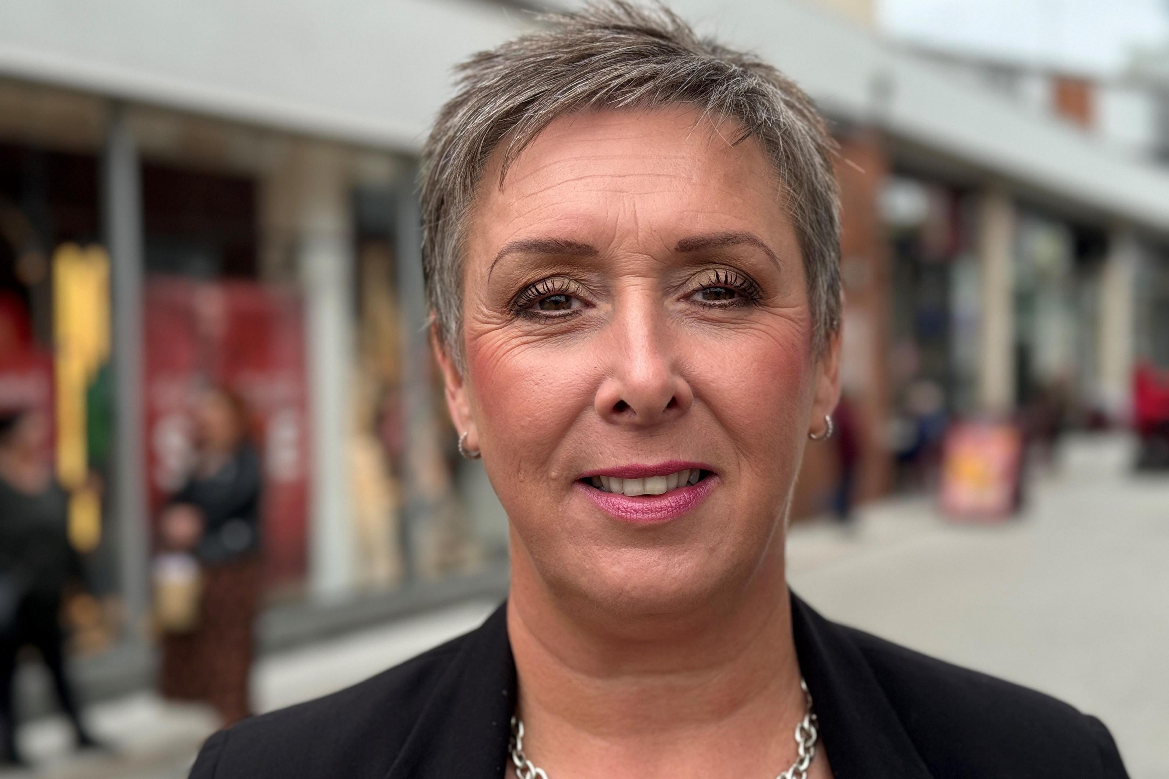 Lisa Fitzgerald stands in front of modern shop fronts at the Pescod Square shopping precinct. She has short hair and wears a smart black jacket and a silver necklace.