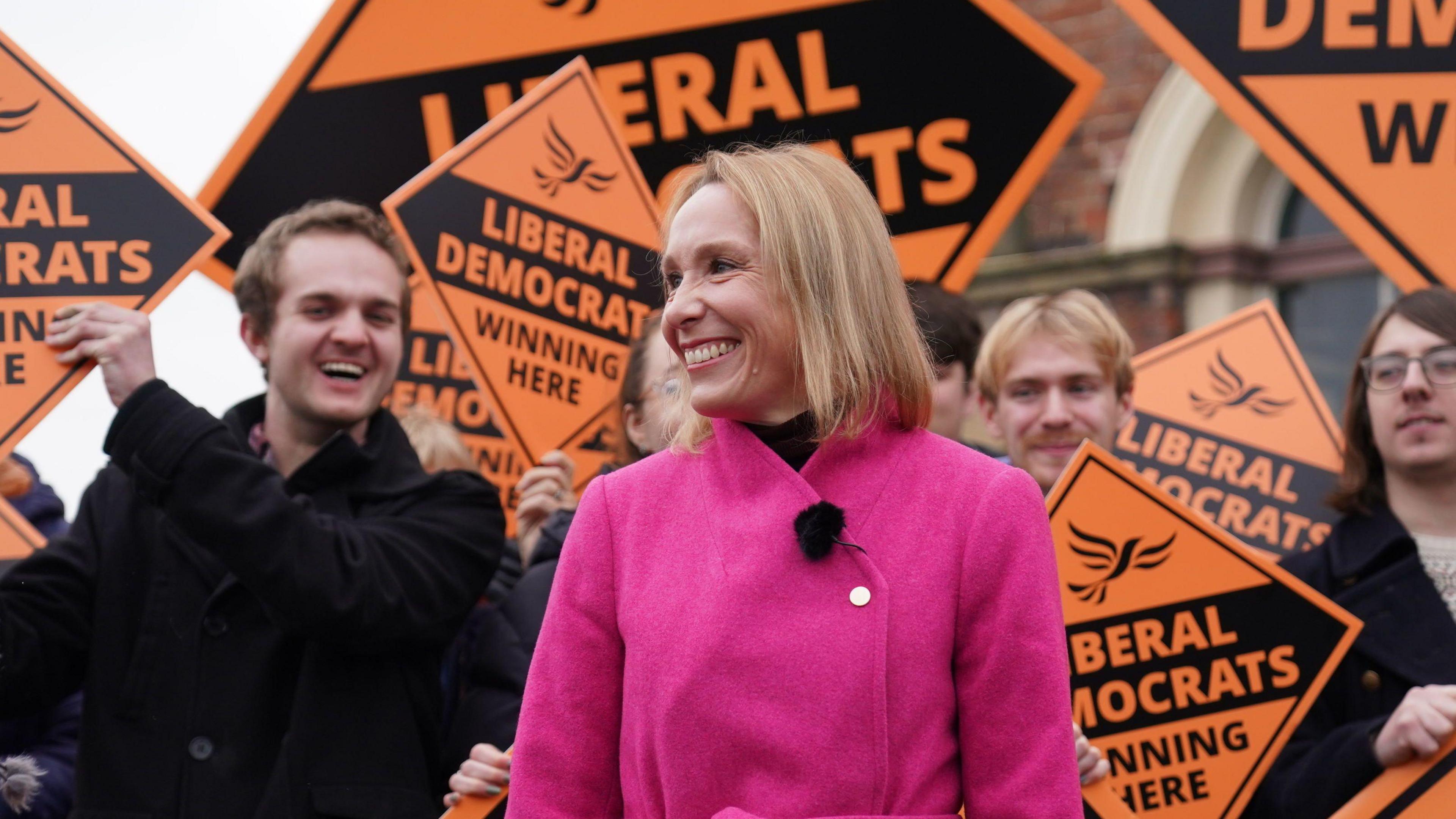 Woman wearing a pink coat smiling in front of orange "Liberal Democrat" campaign boards and happy-looking supporters