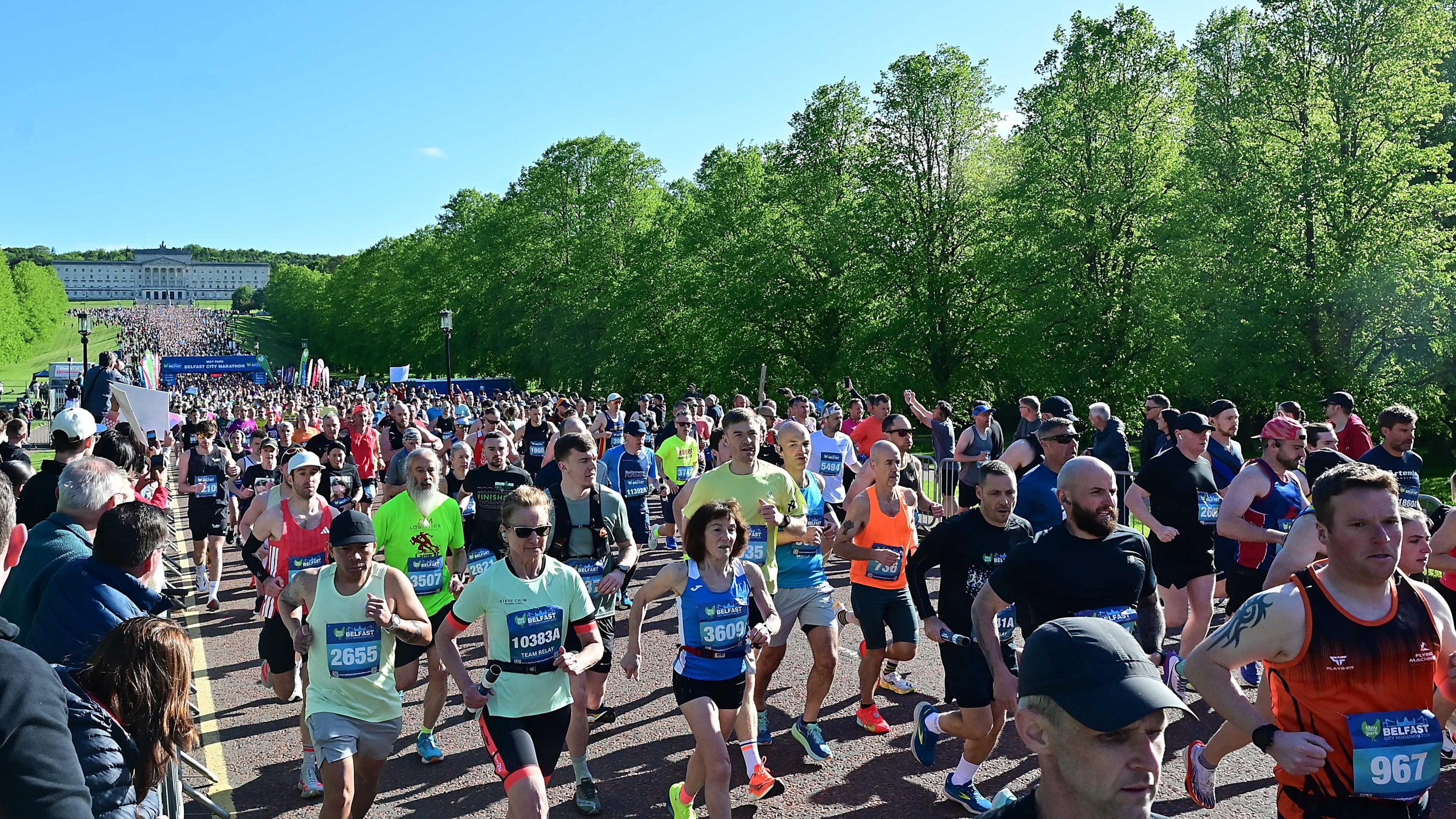 Numerous runners take off from the marathon start line wearing various coloured sportswear. There are spectators along the side of the road and Stormont in the background.