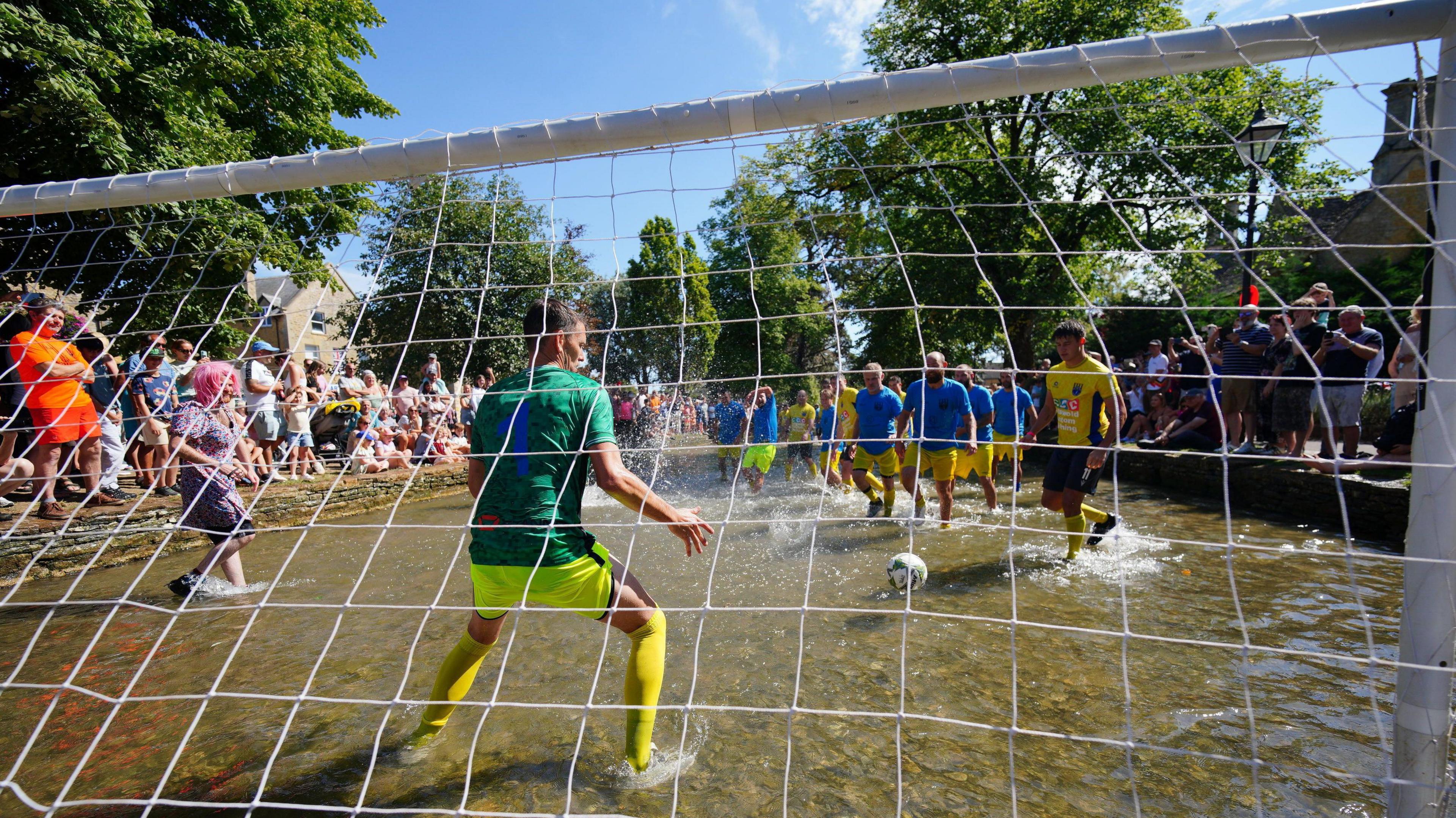 A view from behind the goalpost net looking through at the players approaching, the ball in the river and the goalkeeper prepared. Crowds line the side of the river.