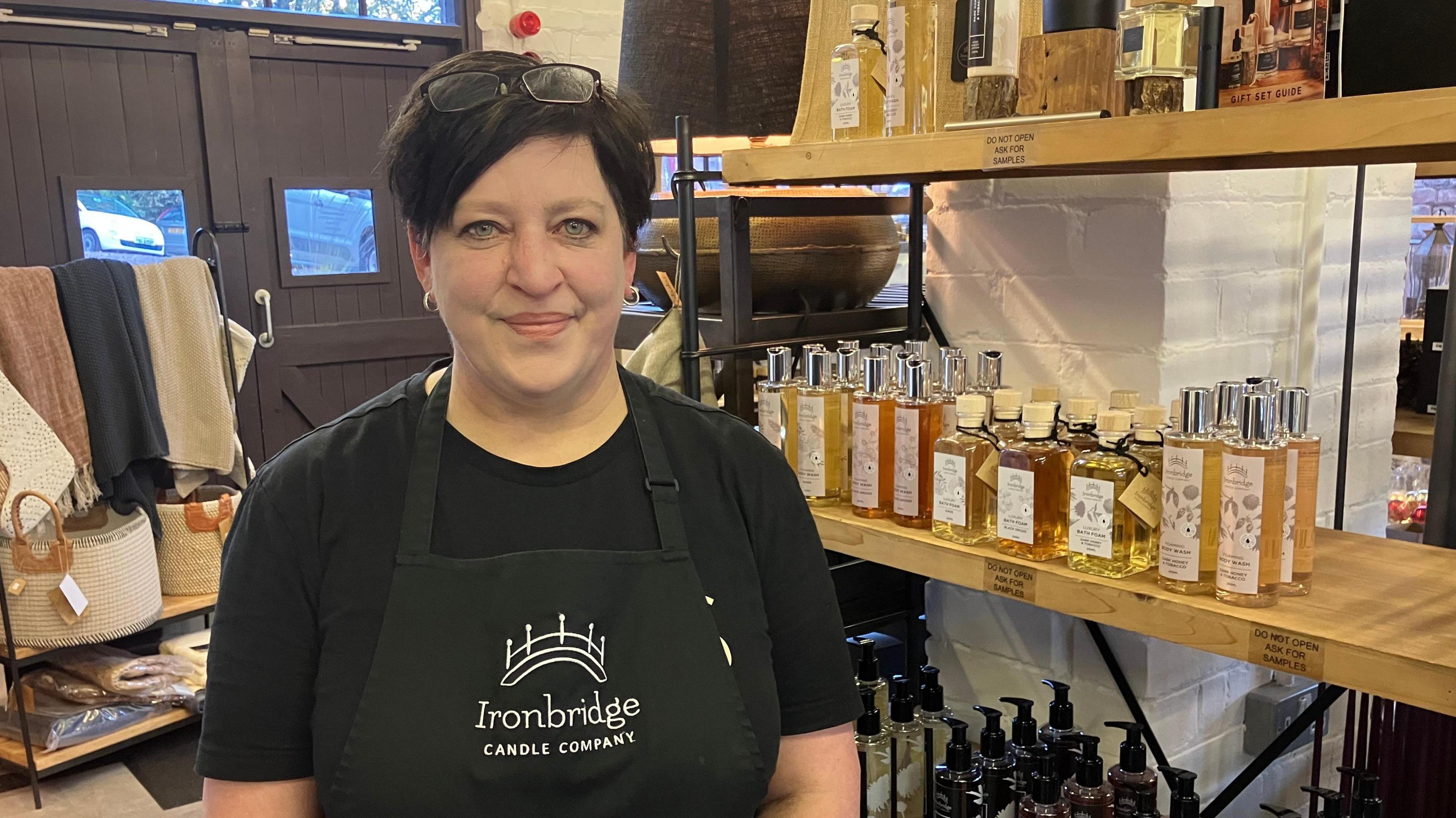 Cherryl Montrose-Matheson standing in her shop wearing a black top and black apron with Ironbridge Candle Company. She is standing in front of a shelf of products and is looking into the camera