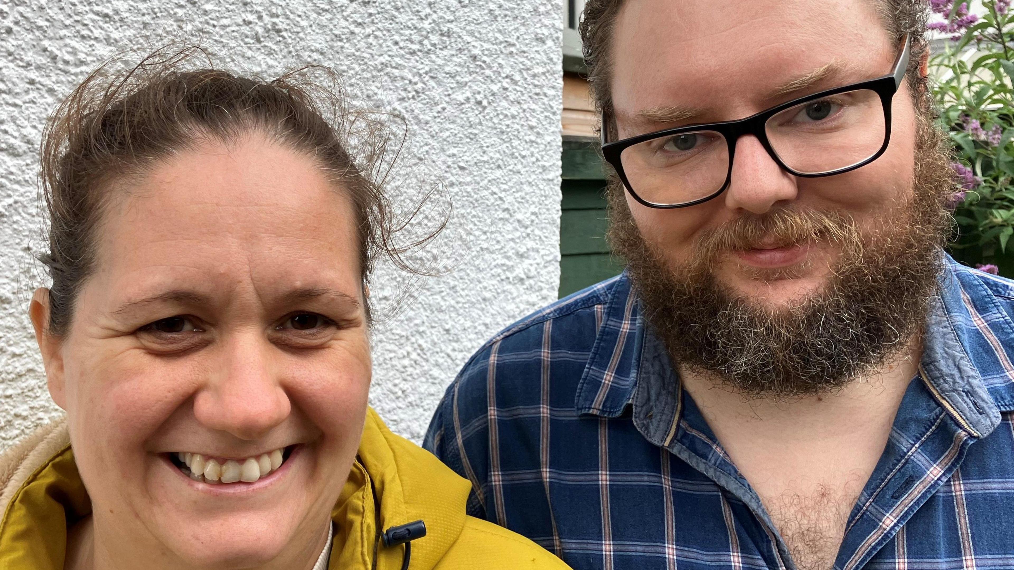 Molly and Gary Casling outside the community larder. Milly has pulled-back brown hair and is wearing a yellow coat. Gary has brown hair and a beard, and is wearing glasses and a blue checked shirt   