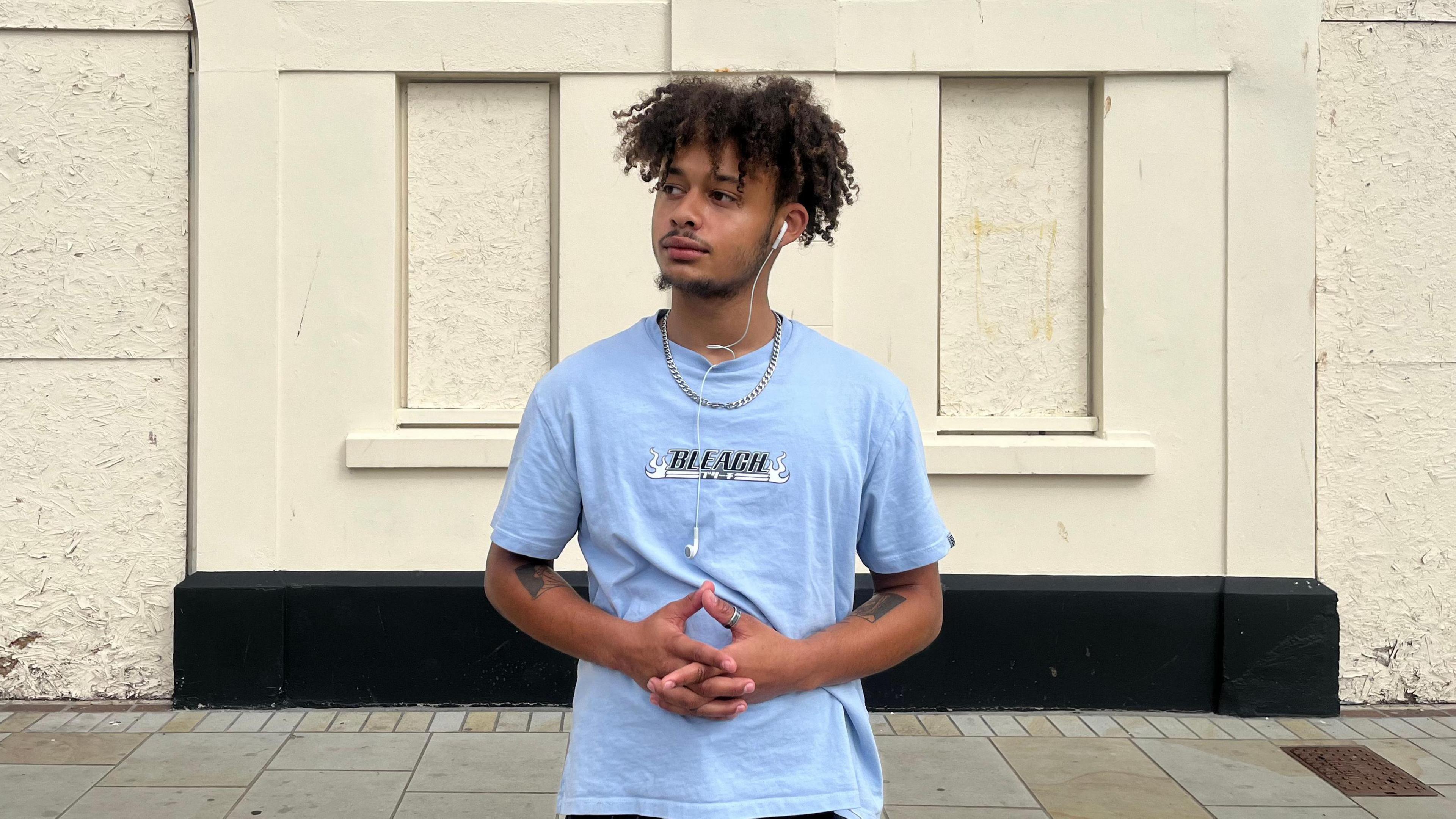 A young man stands in front of a run-down, cream rendered building with his hands folded in front of him. He is looking into the distance and is wearing a light blue T-shirt with the word "Bleach" on it in a black font with white decals, and is wearing a silver ring on one thumb and a silver chain around his neck. He is wearing black jeans and has wired headphones, one of which is hanging out and dangling on his chest.