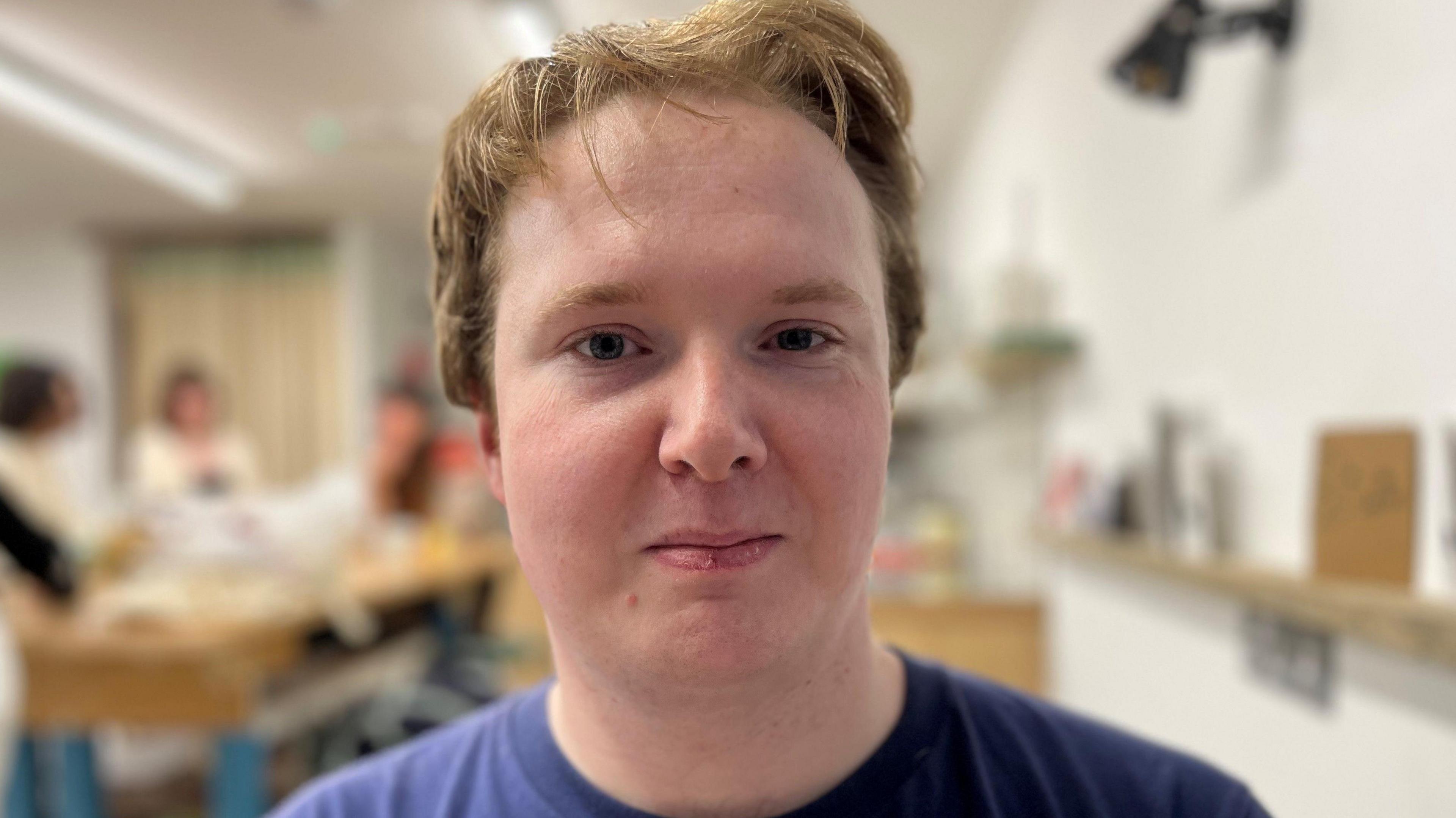 A young man with reddish short hair and brown eyes looking directly at the camera and half smiling, he is also wearing a blue t-shirt