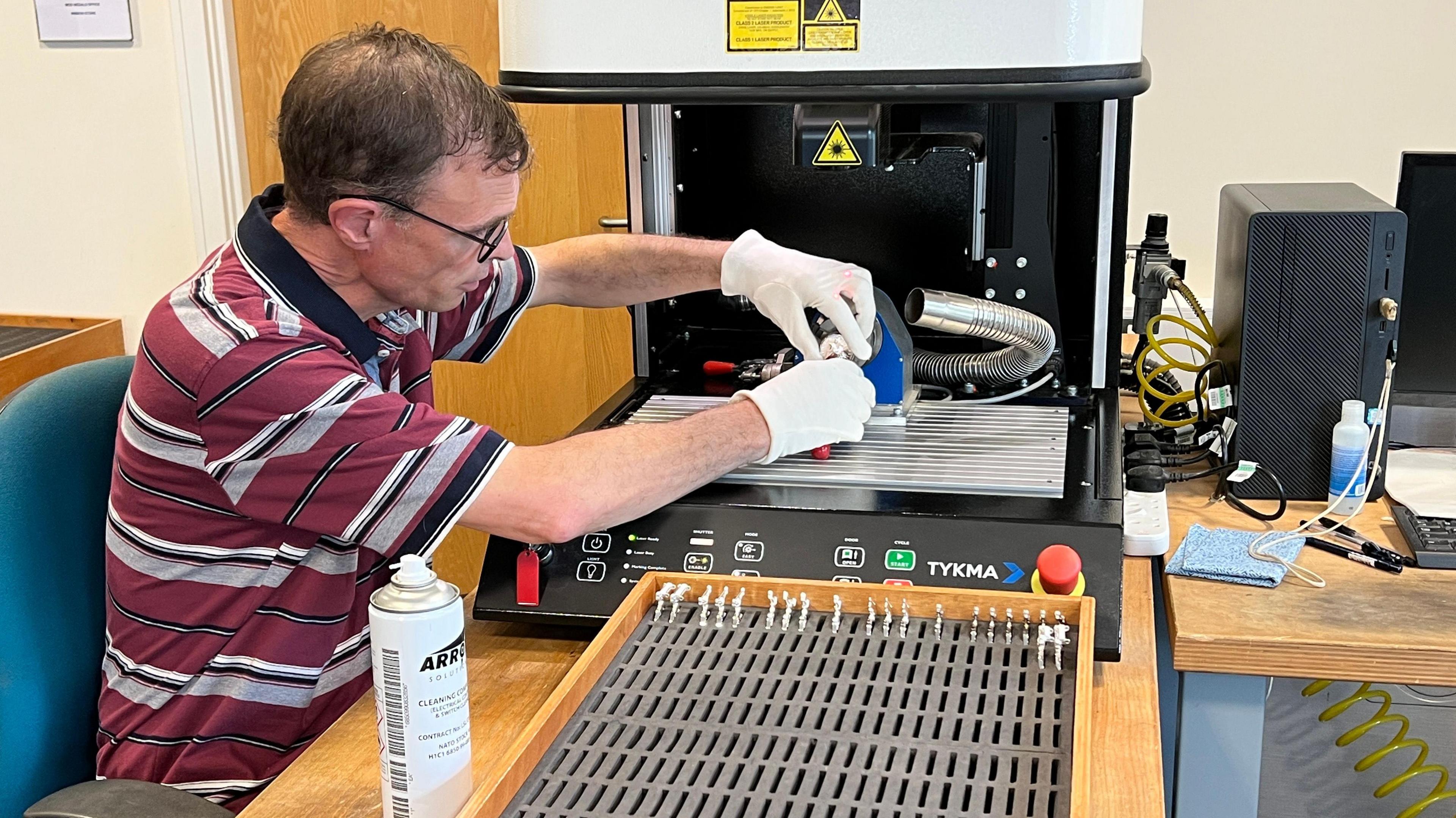 A person wearing a striped shirt and white gloves operates a laser engraving machine, placing a medal inside. A can of ARP spray and a tray of small metal items are visible on the worktable, alongside various tools and equipment.