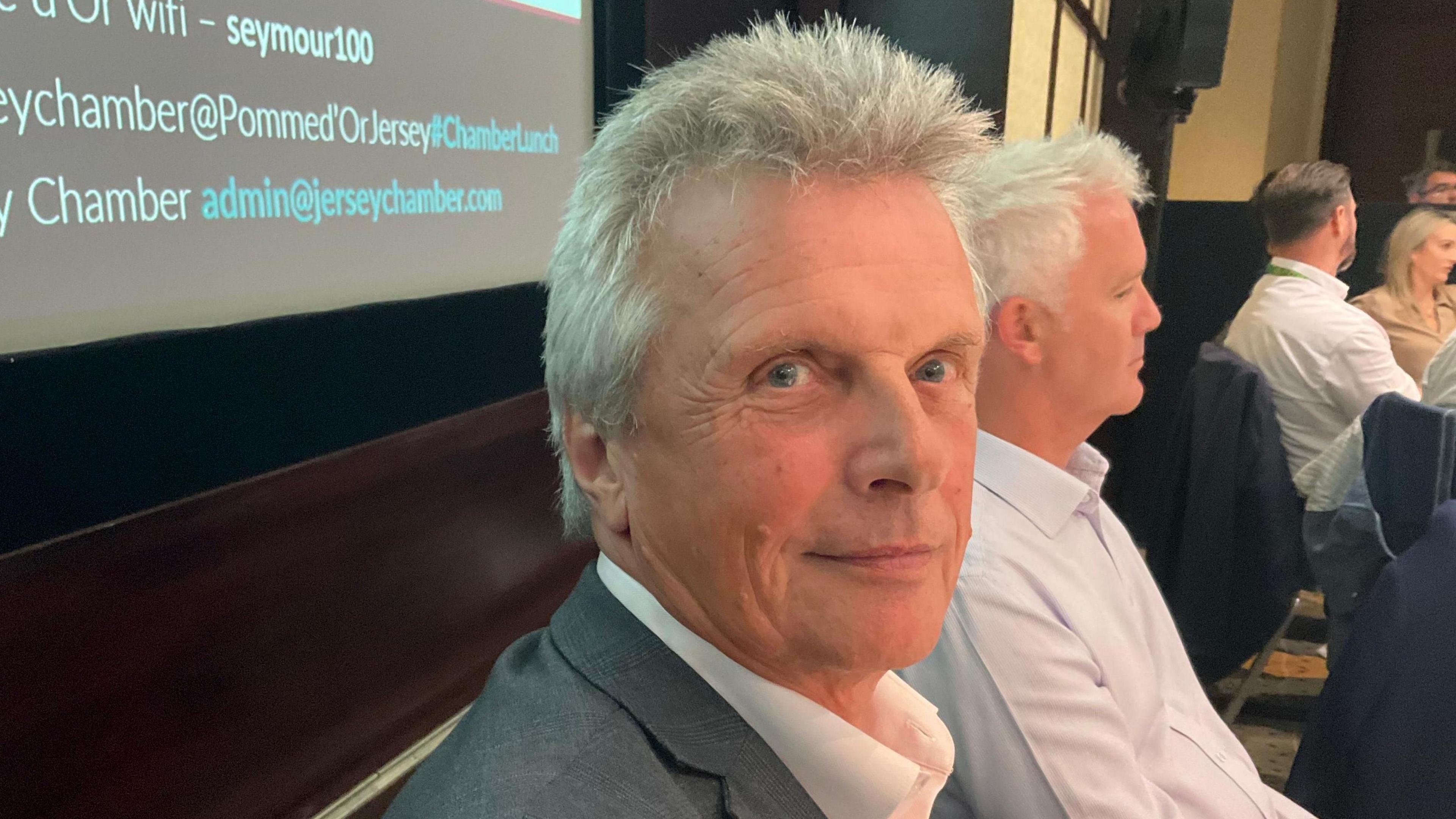 David Seymour sat at a table. He is wearing a grey suit and a white shirt. He has short grey hair. Another man with grey hair is sat next to him looking away front he camera.