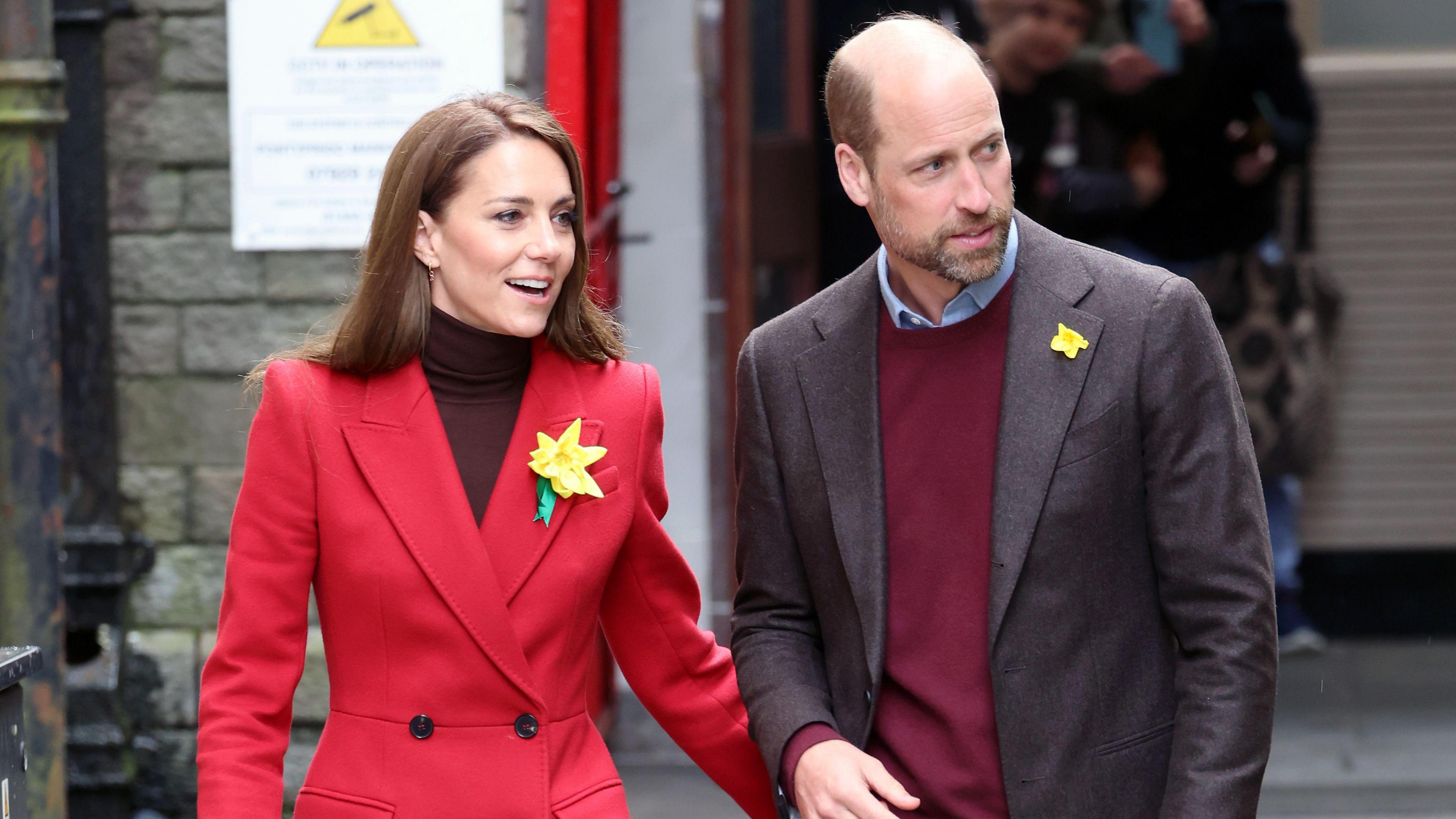 Princess Kate and Prince William wearing formal attire with daffodils attached to their jackets. Kate is wearing a red coat with a turtleneck. He is wearing a suit jacket with a burgundy sweatshirt underneath.