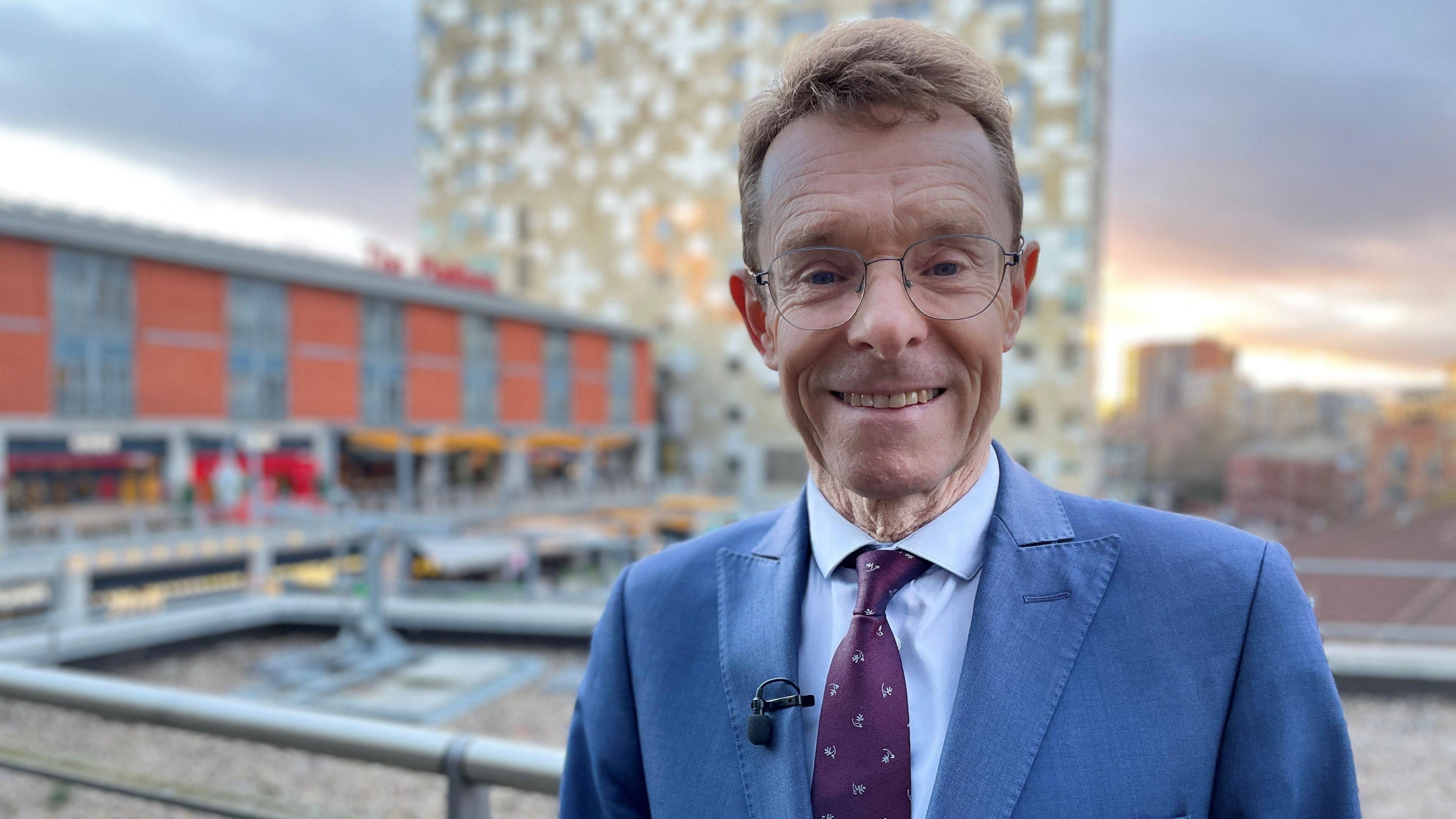 A man with brown hair and large wire-framed glasses in a blue suit and purple tie with a big smile on his face. In the background there is a blurred view of the canalside in Birmingham with metal railings in front of a row of bars and a tall multi-coloured building in the distance