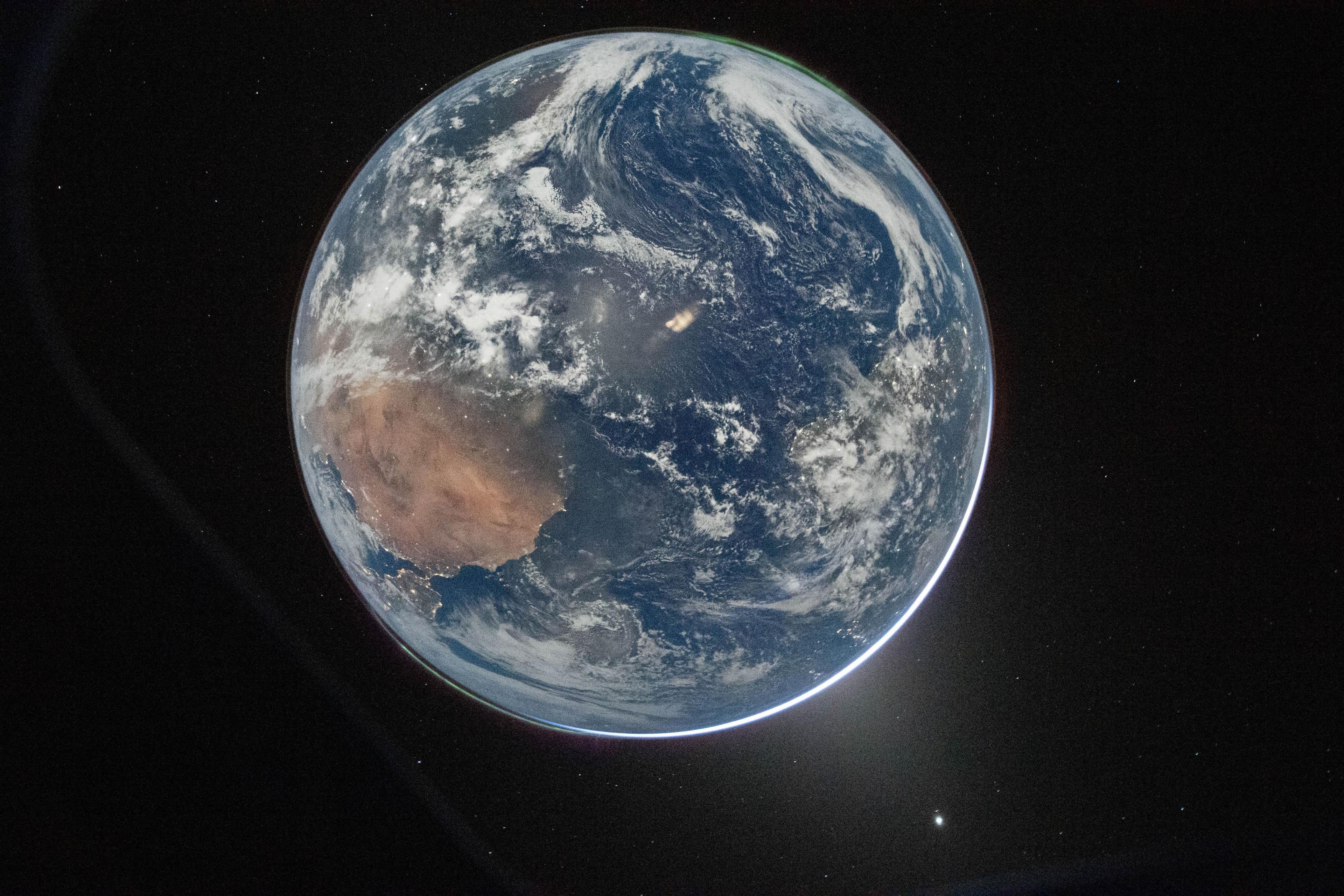A picture of the Earth from space, which centres the planet against the dark background of space. It's a round blue planet. Clouds can be seen and a thin green aurora at the top.