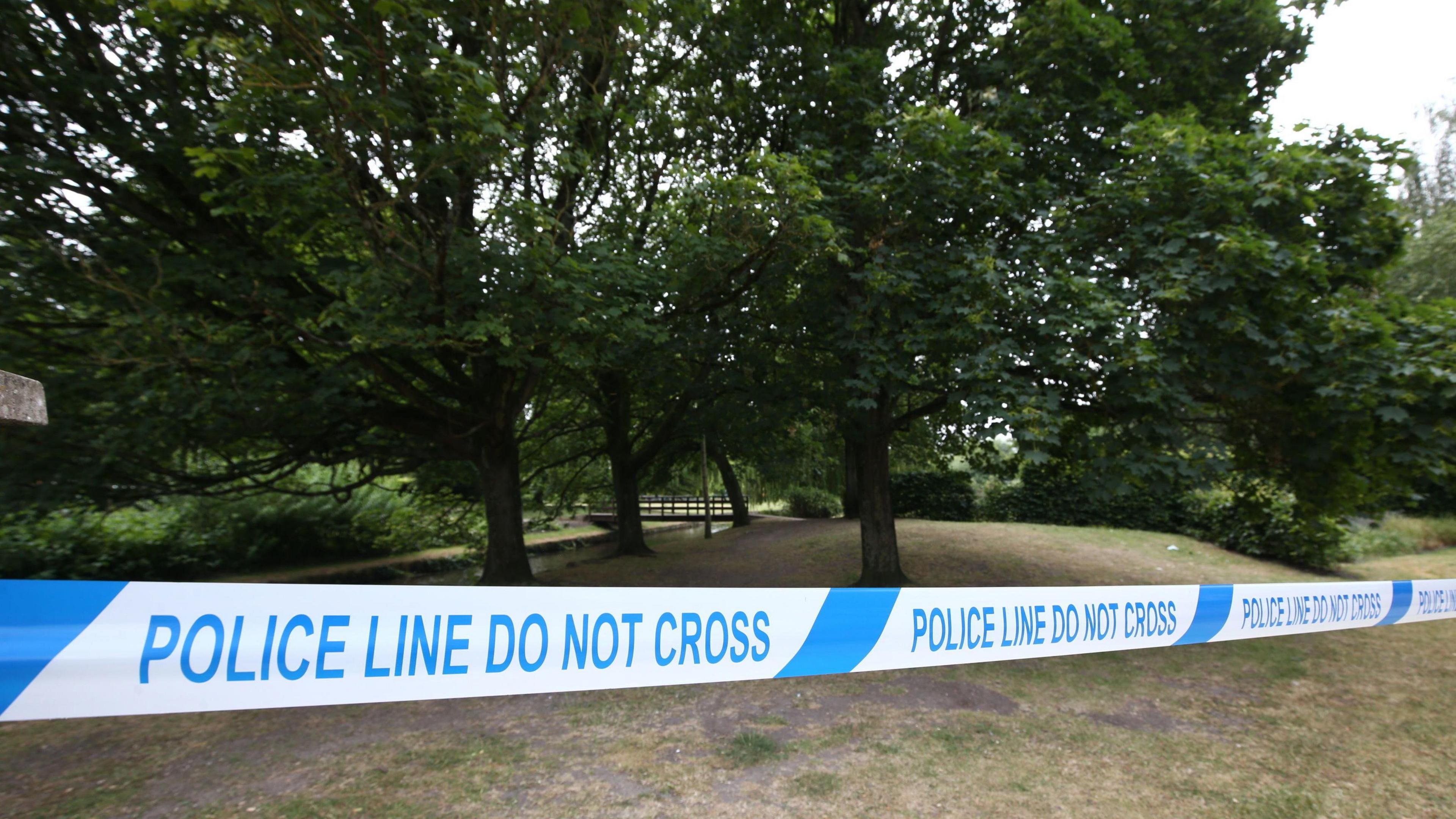 A "do not cross" police line in a park with trees behind