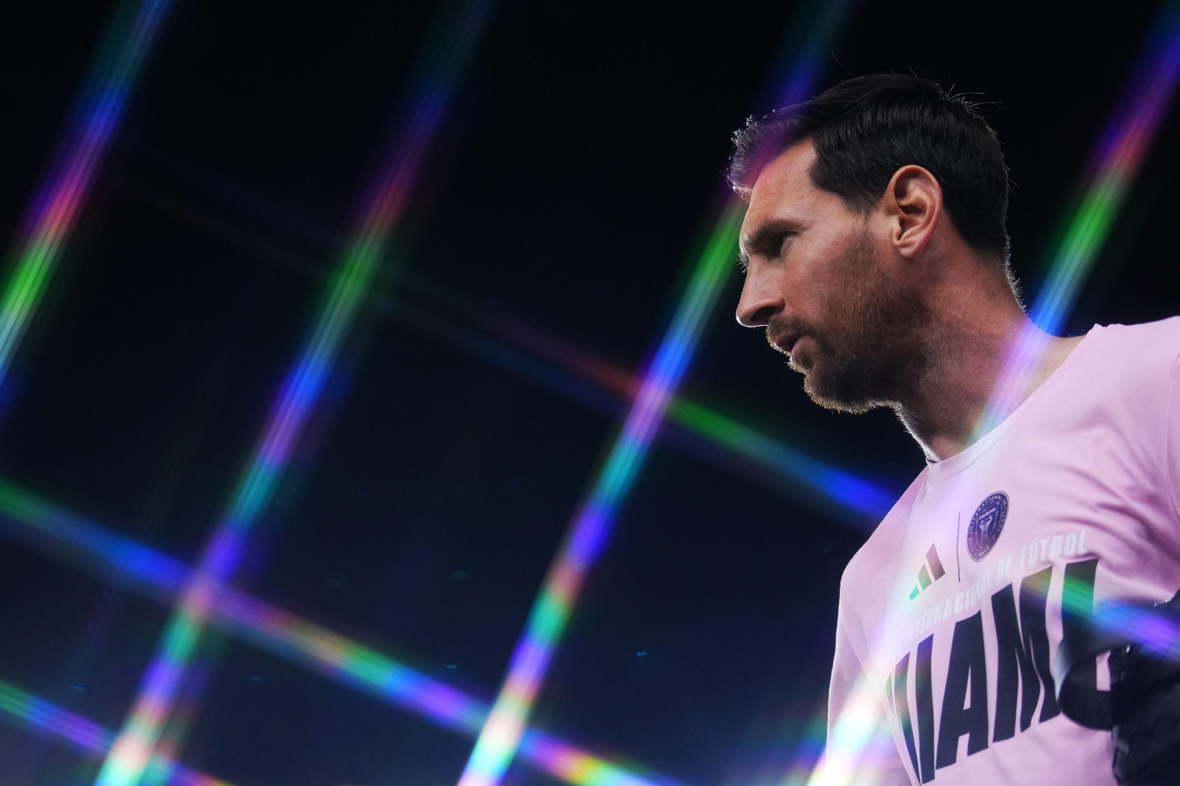 Lionel Messi wearing a white sports jersey with the word ‘MIAMI’ visible, standing under bright stadium lights that create colorful diagonal beams across a dark background, giving the scene a dramatic and vibrant atmosphere.
