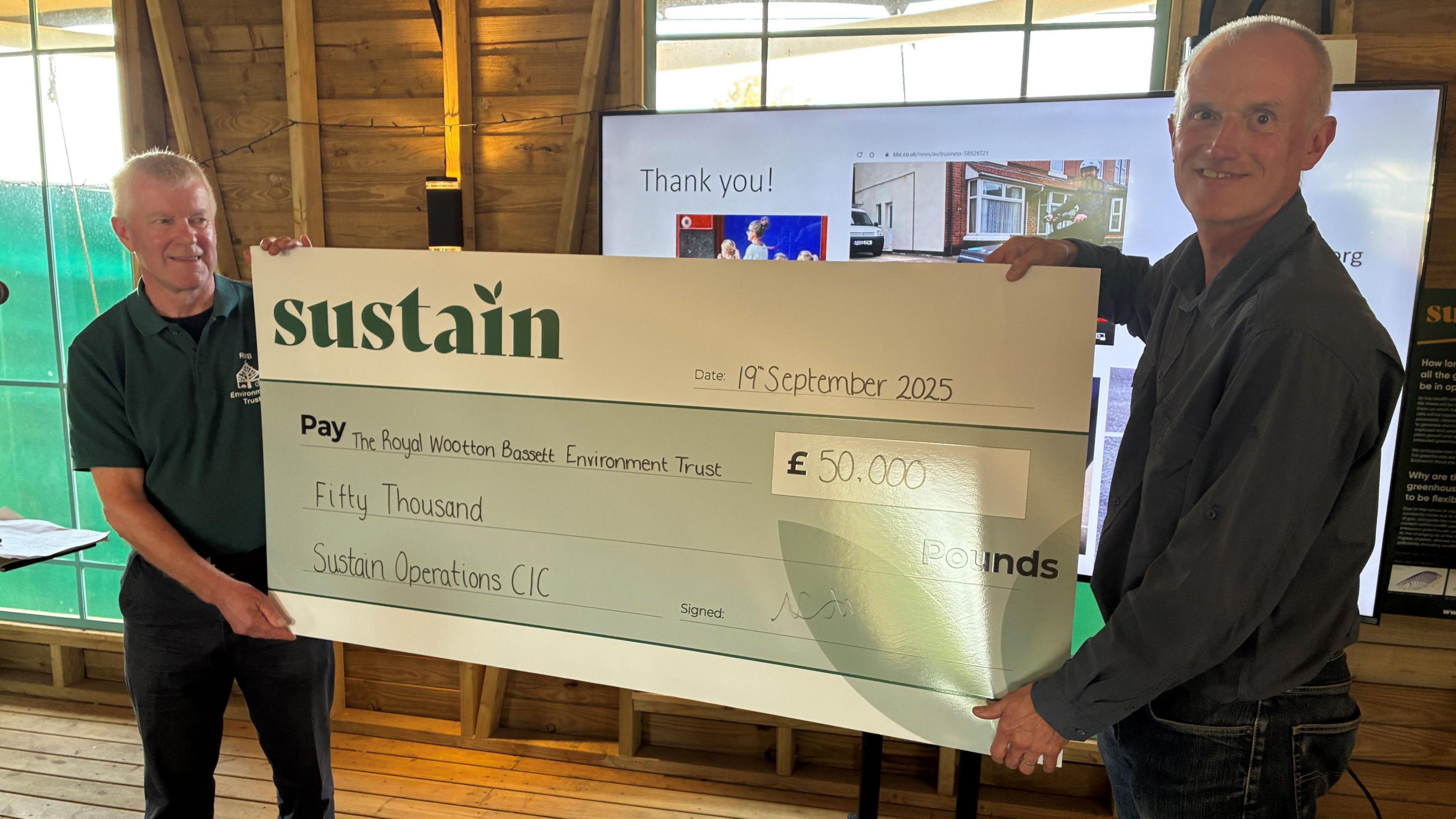 Two men stand smiling either side of a giant cheque for £50,000. They are standing in a wooden building with a television screen behind them. The top of the cheque says "Sustain".