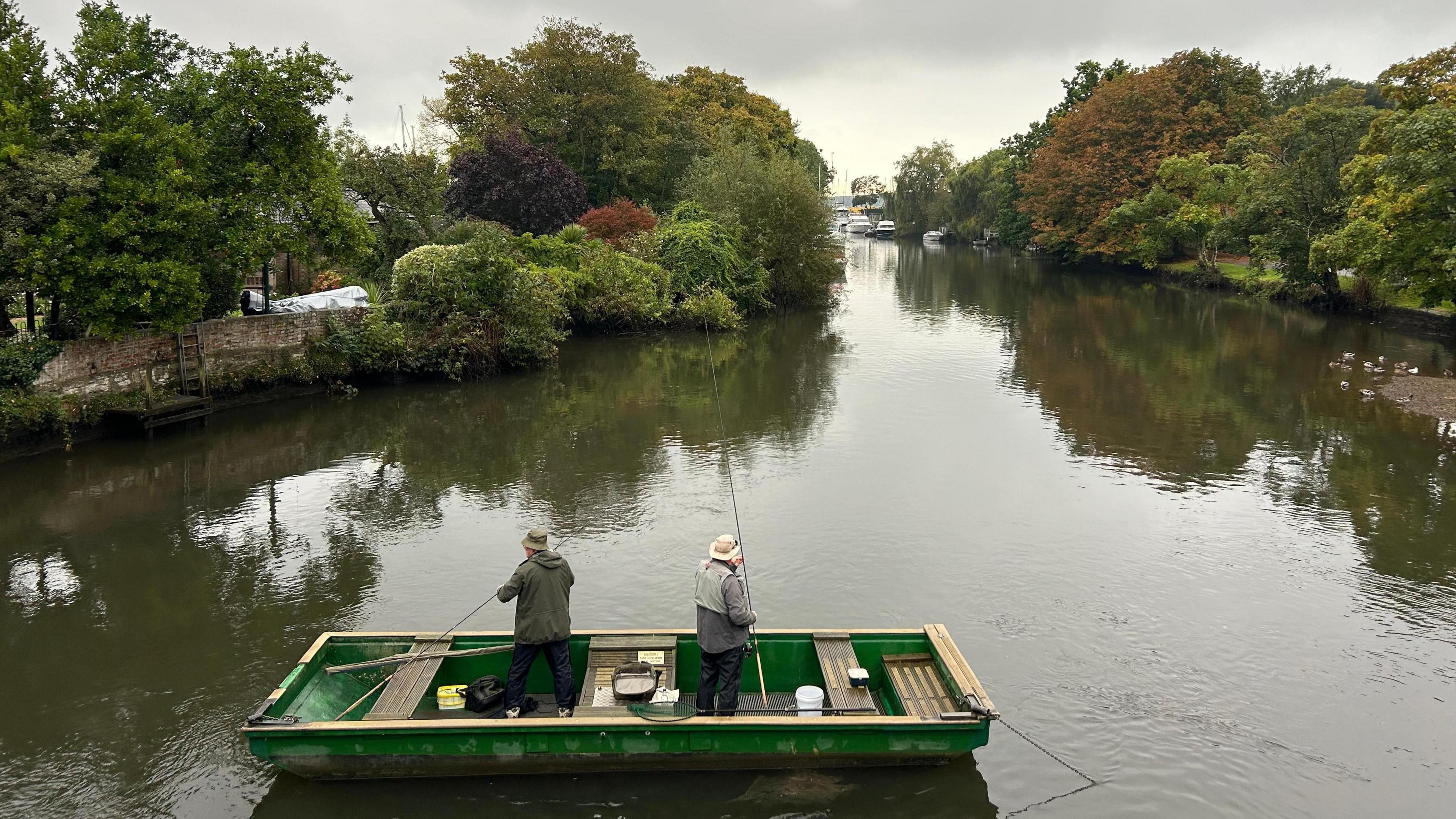 Two fishermen in a green boat on a dark river are both holding fishing rods. They wear green jackets and bucket hats. Each river bank has green trees. The sky overhead is grey.