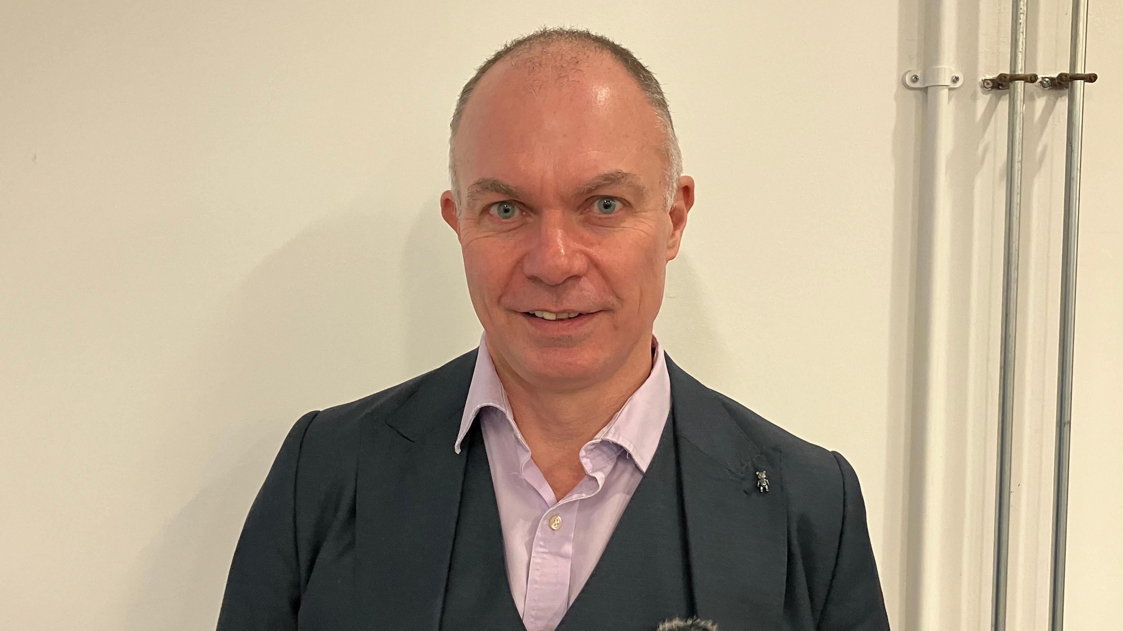 A man in a grey suit , lilac shirt and stood in front of a white wall 