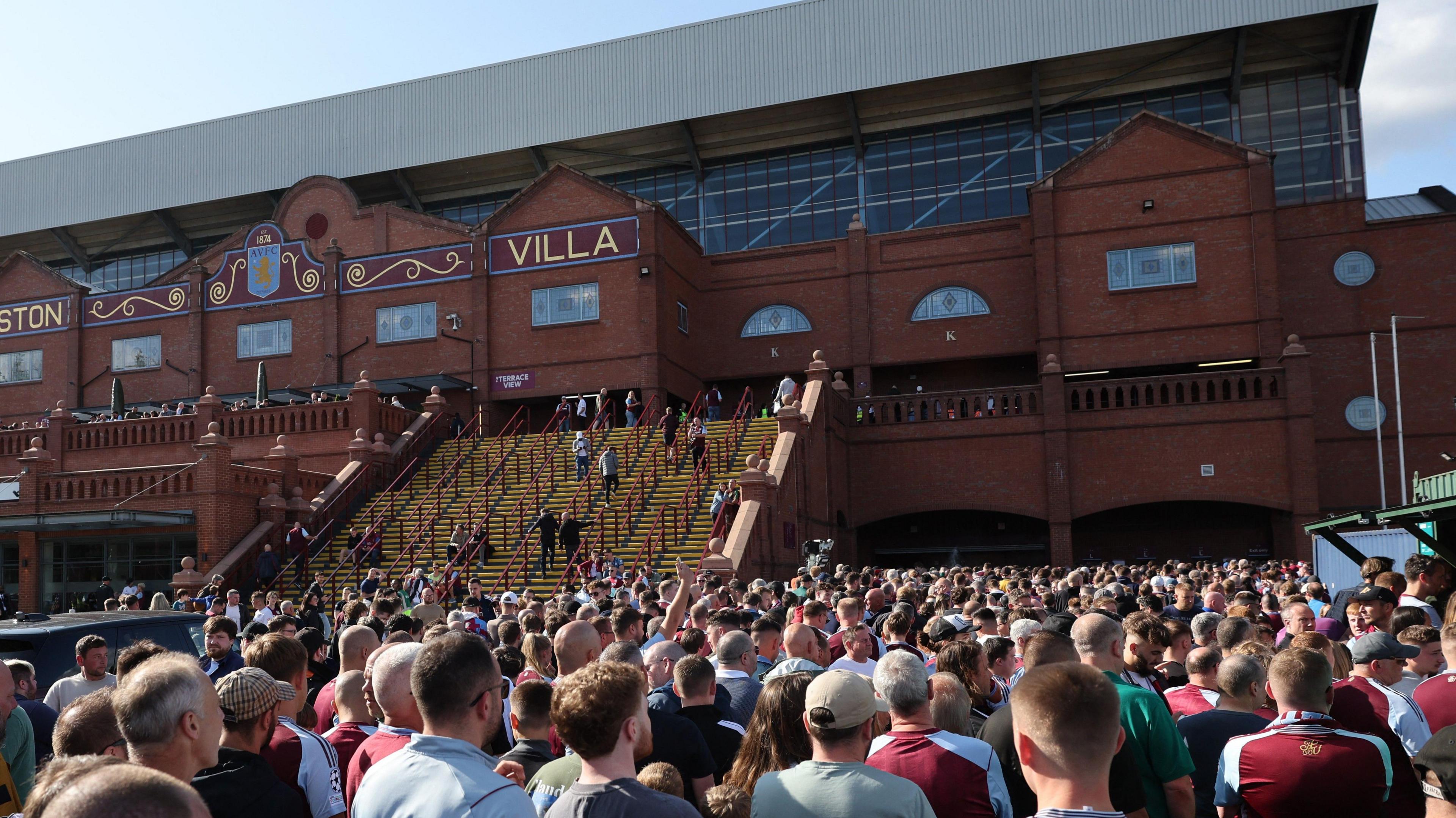 A crowd of Aston Villa fans, all facing away from the camera, queue to get into the Holte End. 