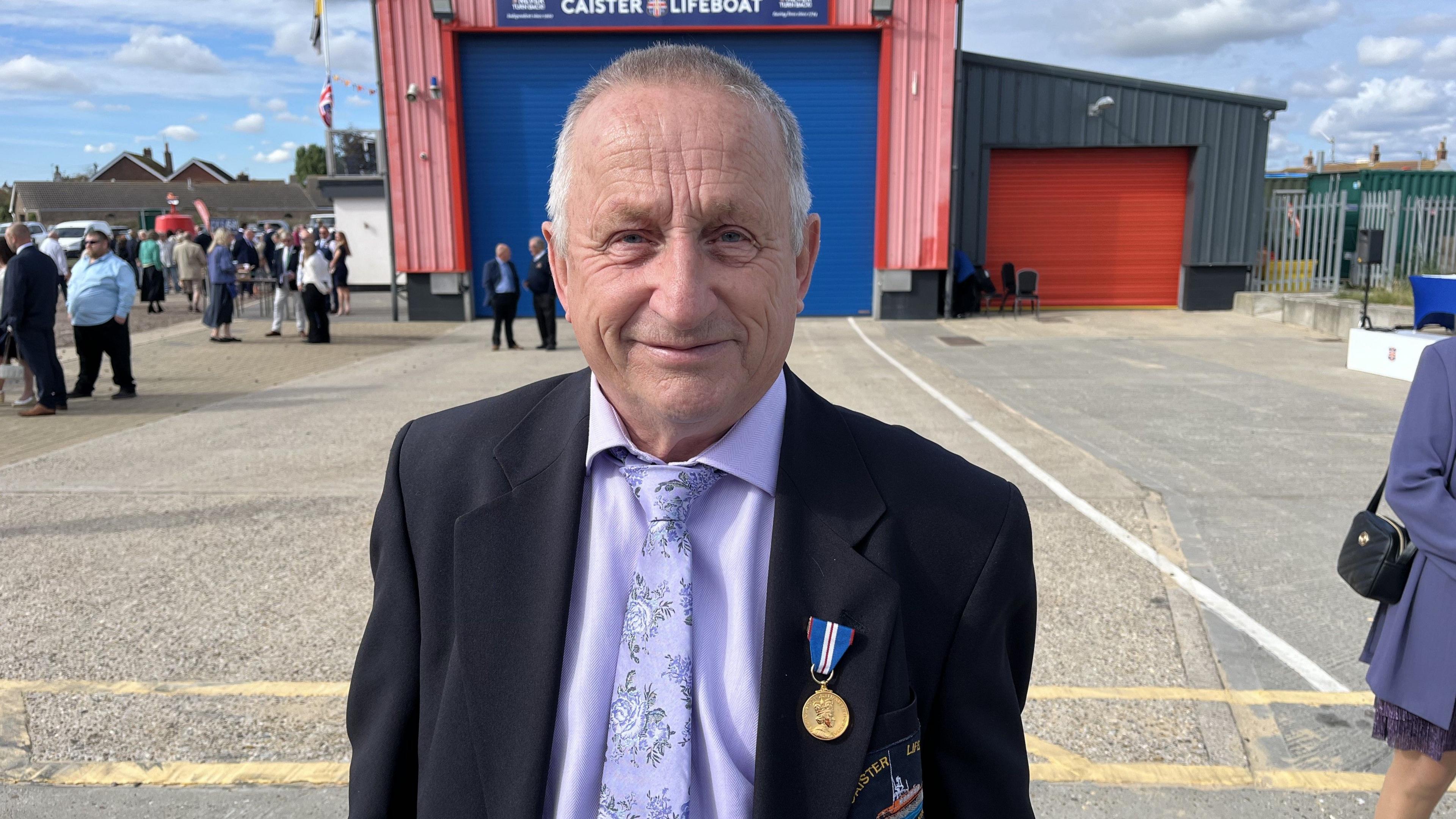 Dick THurlow is looking directly in the camera and is wearing a lilac shirt and tie and a blazer with a decorated medal on the lapel