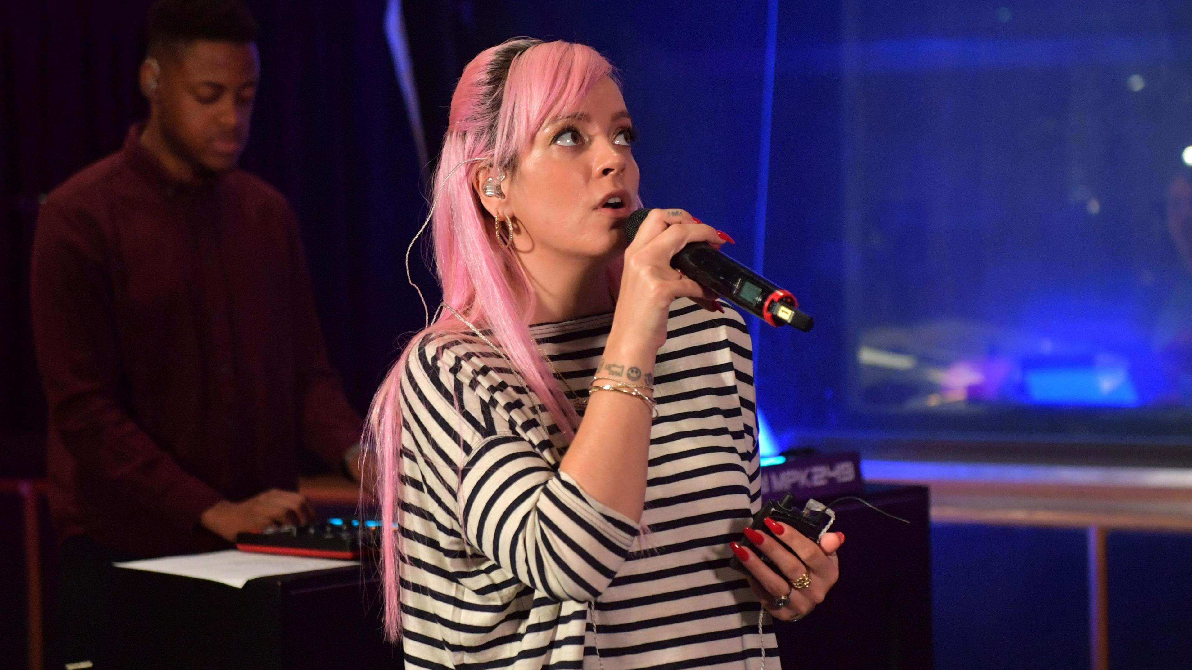 Lily Allen performing. She has long pink hair and is wearing a black and white top.