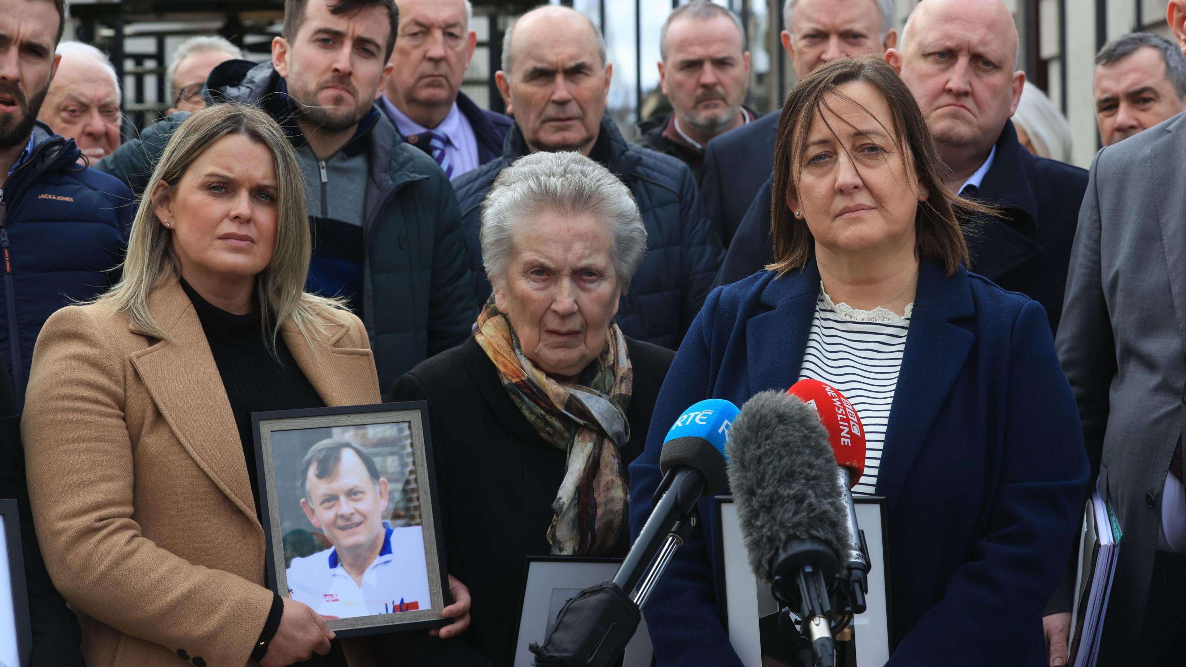 Three women holding photographs of Sean Brown standing at microphones outside. A group of about 10 men are standing behind the.