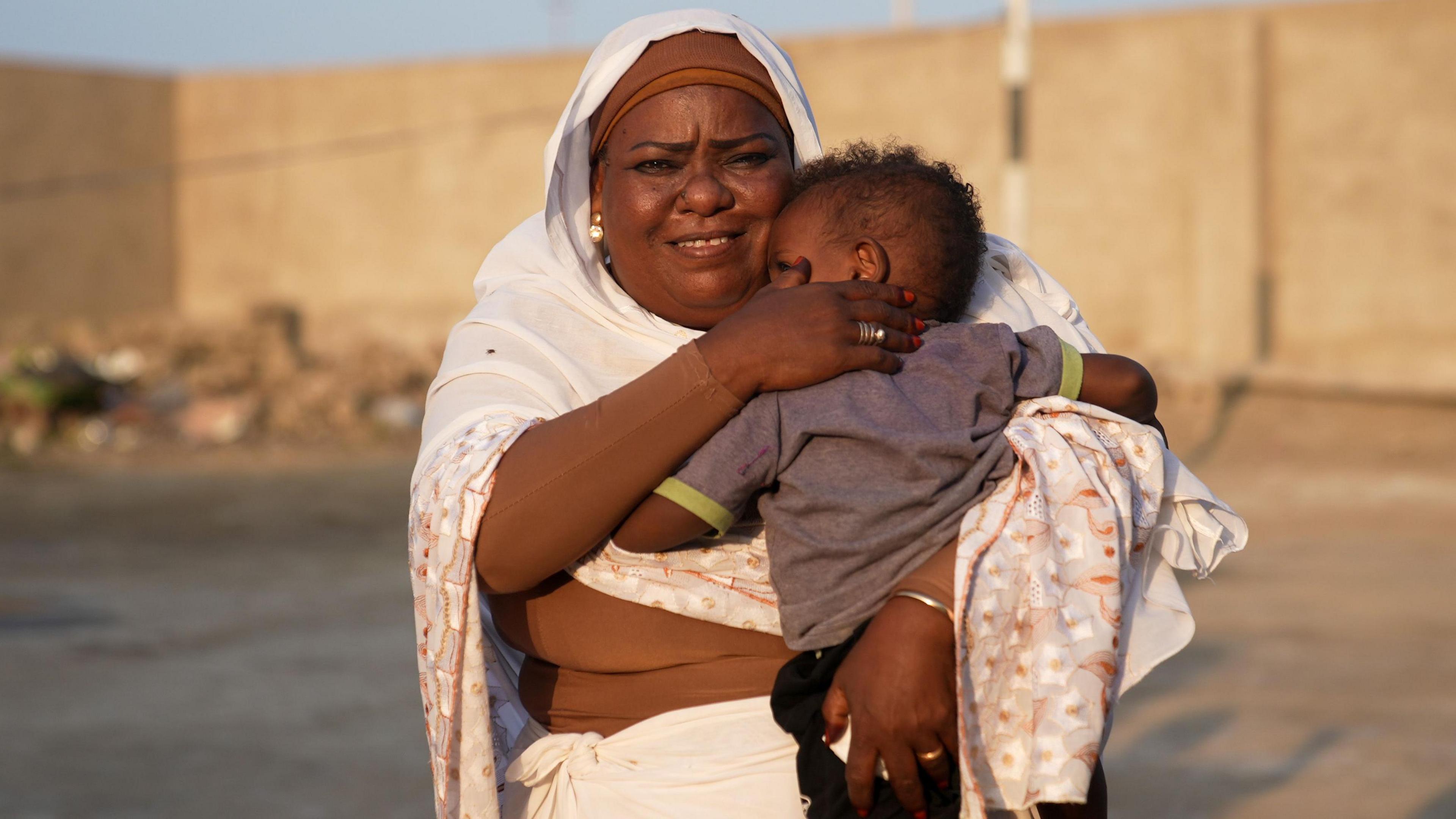 Mama Nour in Port Sudan stands outside looking at the camera clutching a child to her