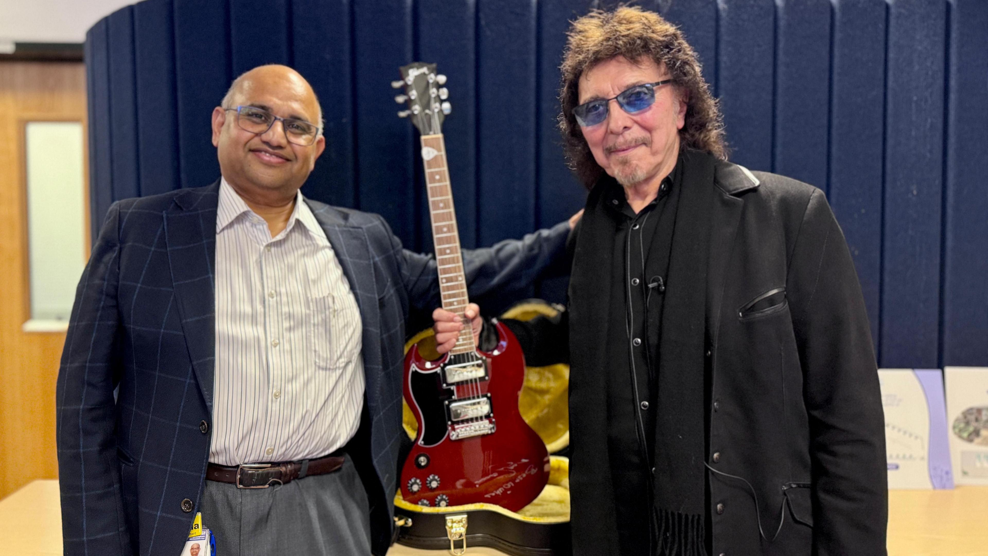 Shankara Paneesha and Tony Iommi are standing together in the hospital unit, holding the guitar between them. Mr Paneesha is wearing a shirt and jacket, and Iommi is dressed in black.