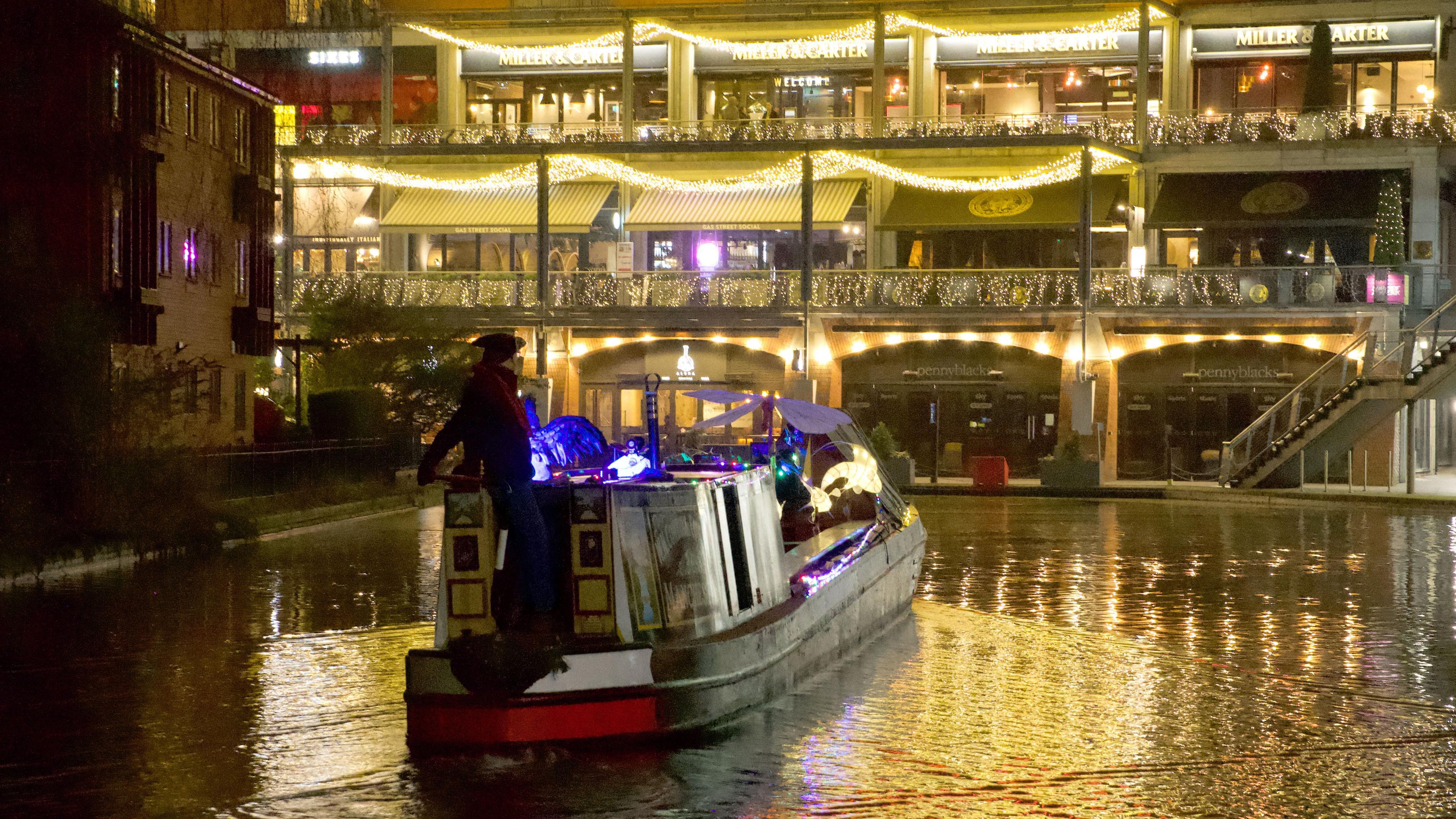 A narrowboat with colour lights on board. The vessel is heading towards a series of golden-lit restaurants across three levels at the Mailbox in Birmingham