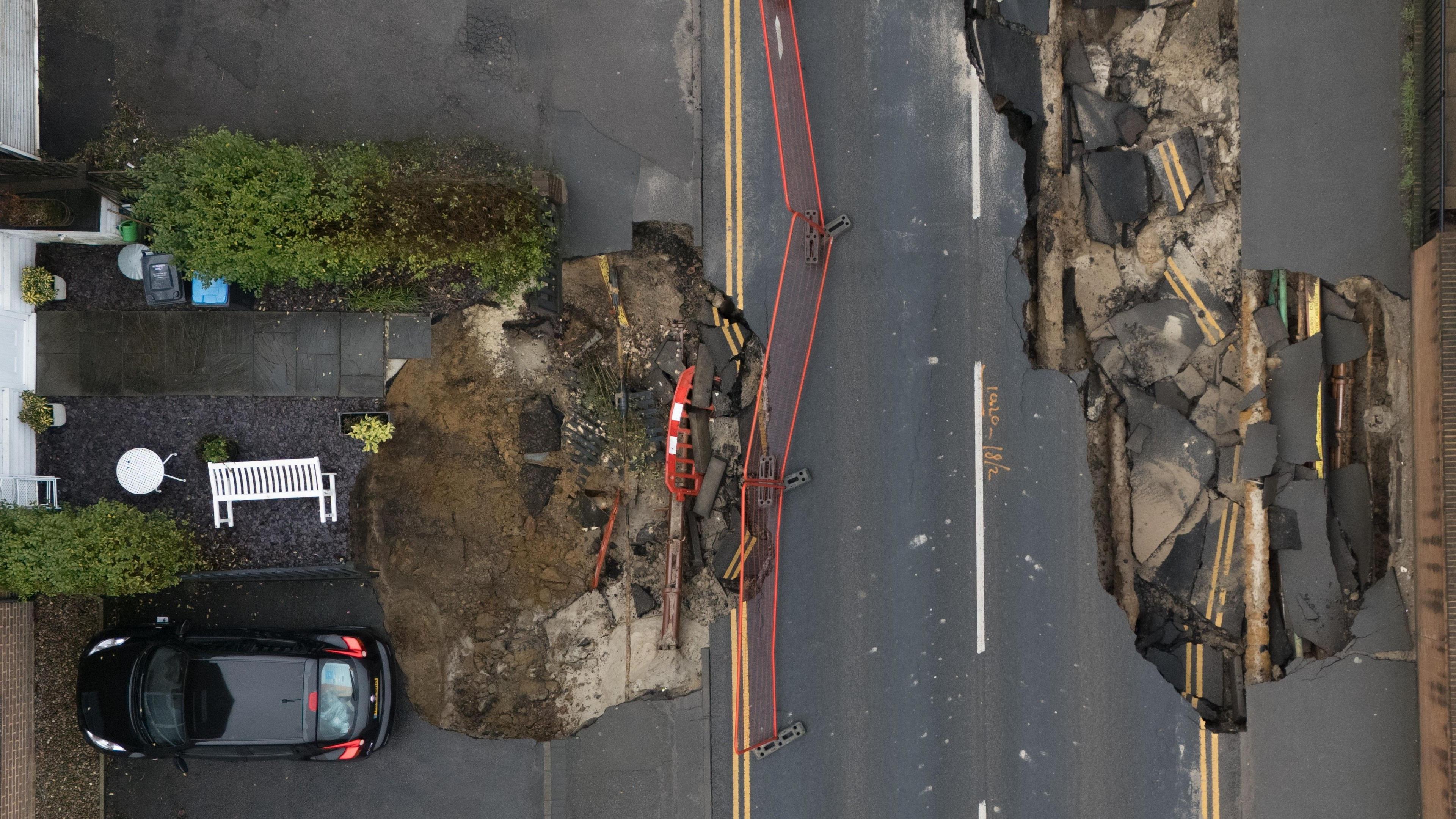 An aerial view of two sinkholes on a residential road. There is a car on the left which appears to be parked right on the border of one sinkhole.