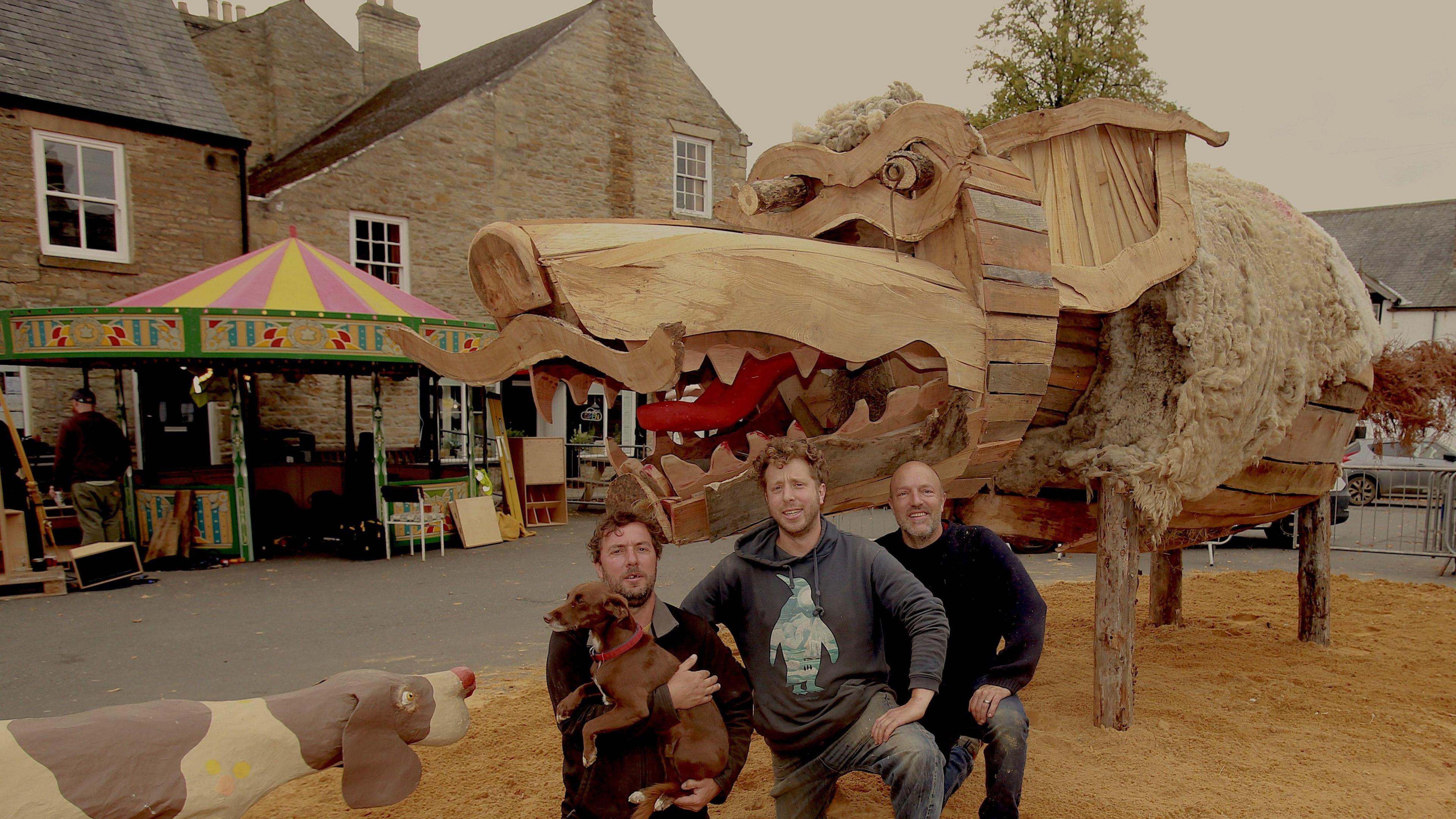 Three men kneeling in front of the large wooden structure of the wolf. They are smiling and the man on the left is holding a small, brown dog. The sculpture has large sharp teeth and a red tongue and is covered in what looks like wool. Stone buildings can be seen in the background.