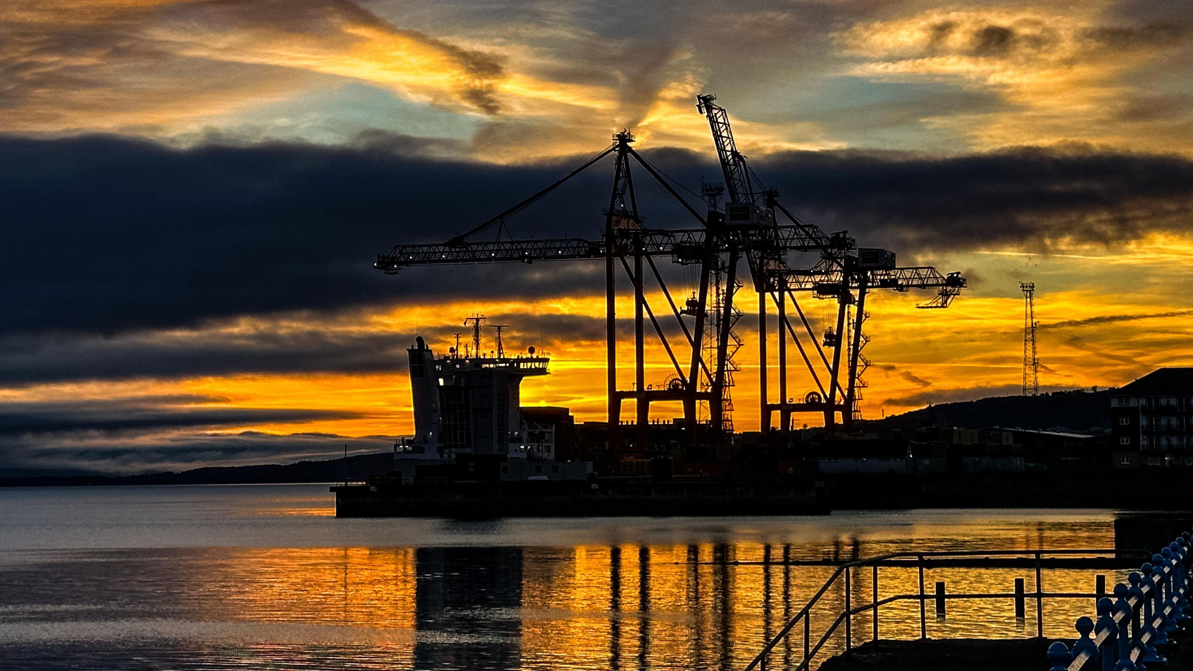 Sunrise over the River Clyde as seen from Greenock. The sky is a mixture of blue, dark , orange and yellow. The sun can almost be seen behind a crane in the background of the picture. Closer to the front, three other cranes, which are dark in colour, hang over the water, which is still.