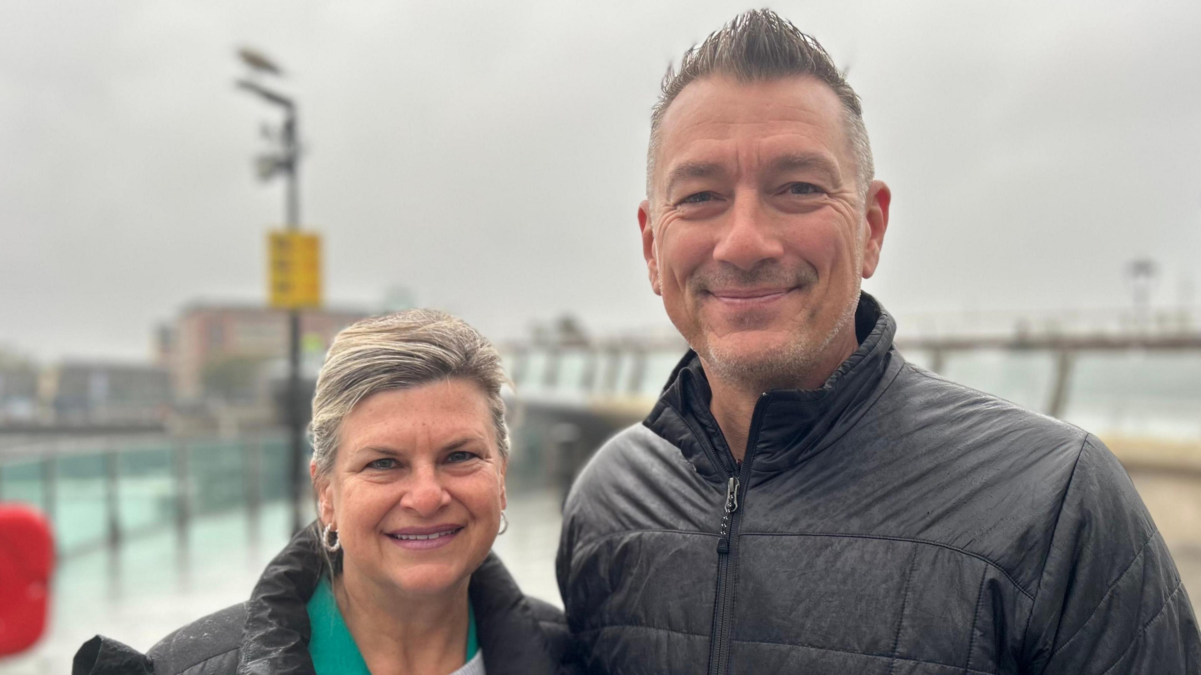 On the left, a long haired blonde woman wearing a black coat and a green top. On the right, a tall dark haired man wearing a black puffy coat. The background is blurred, but it looks like a wet and cloudy day.