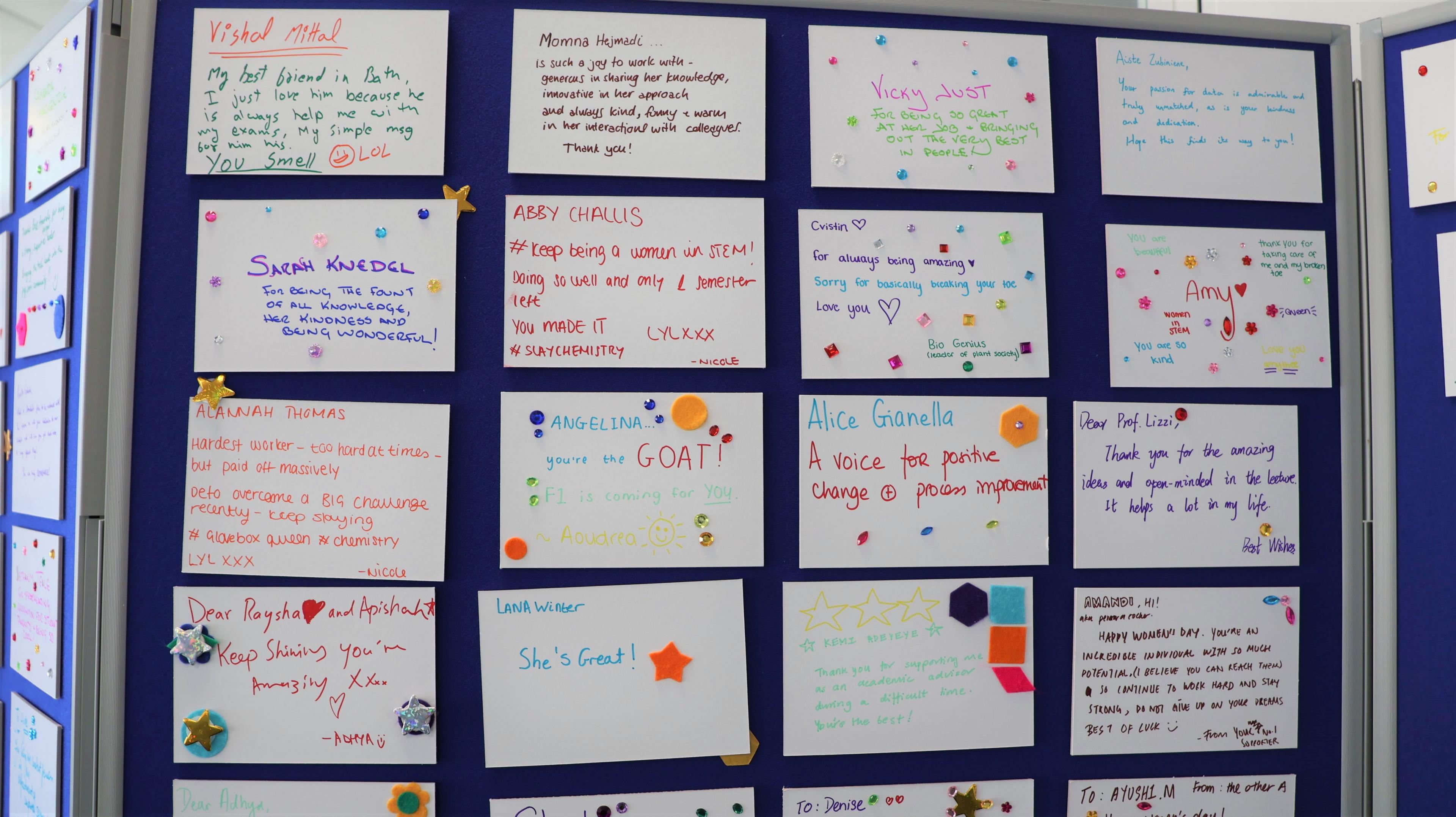 A close up of the wall of nominations displayed on a blue notice board. One says 'keep being a woman in STEM', another says 'a voice for positive change', and another says 'thank you for the amazing ideas'. They are all written in colourful pen, with hearts and kisses drawn on them. 